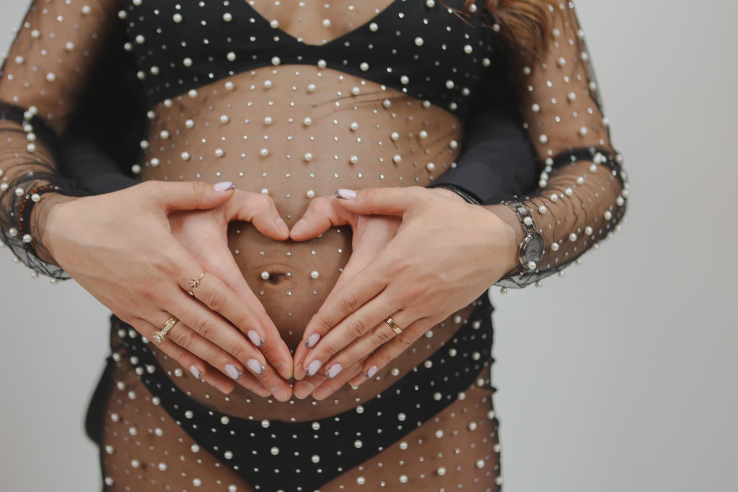 Pregnant woman in sheer black pearl dress making a heart shape with her hands over her baby bump.