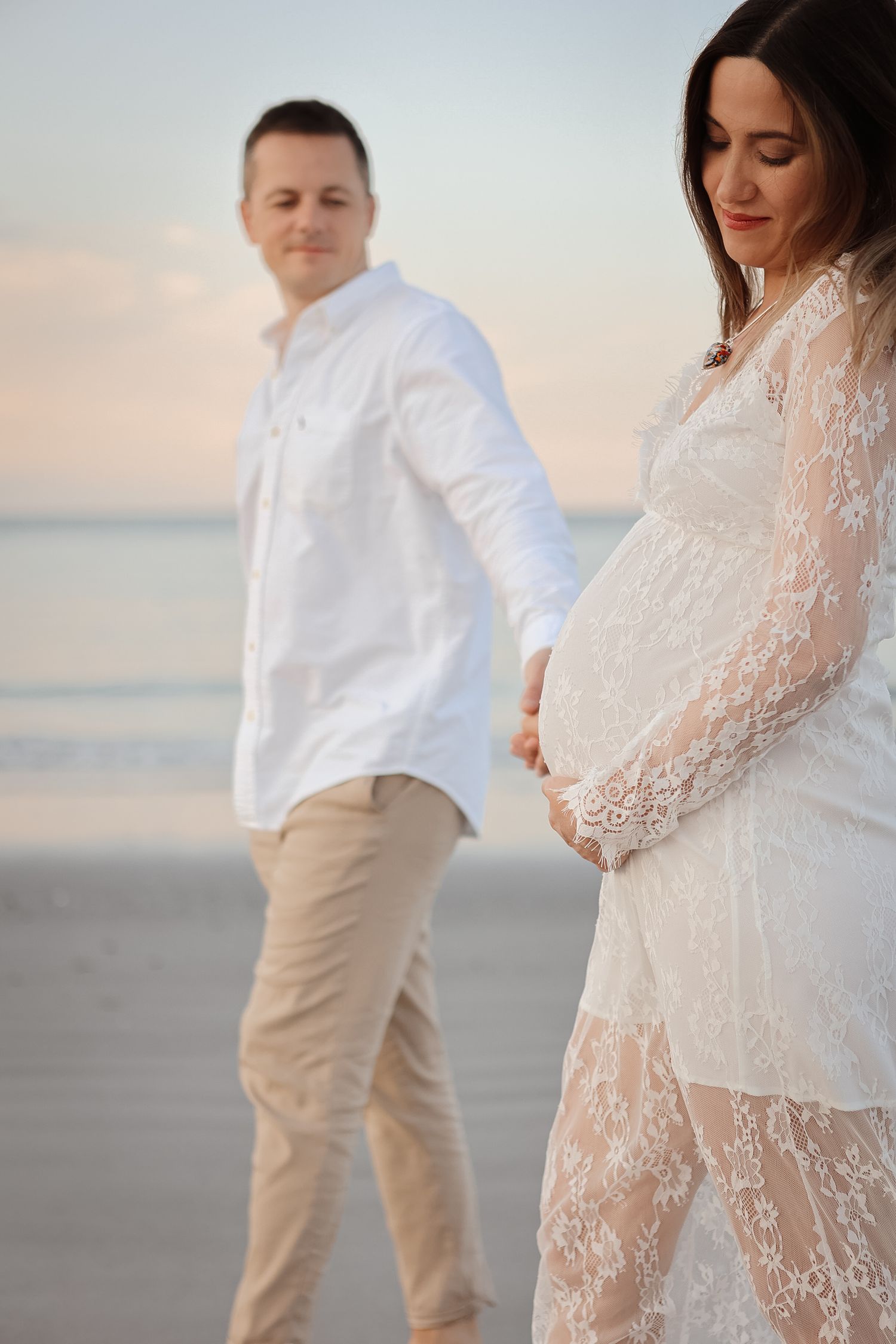 Pregnant woman in white lace dress on beach at sunset with husband standing behind her.