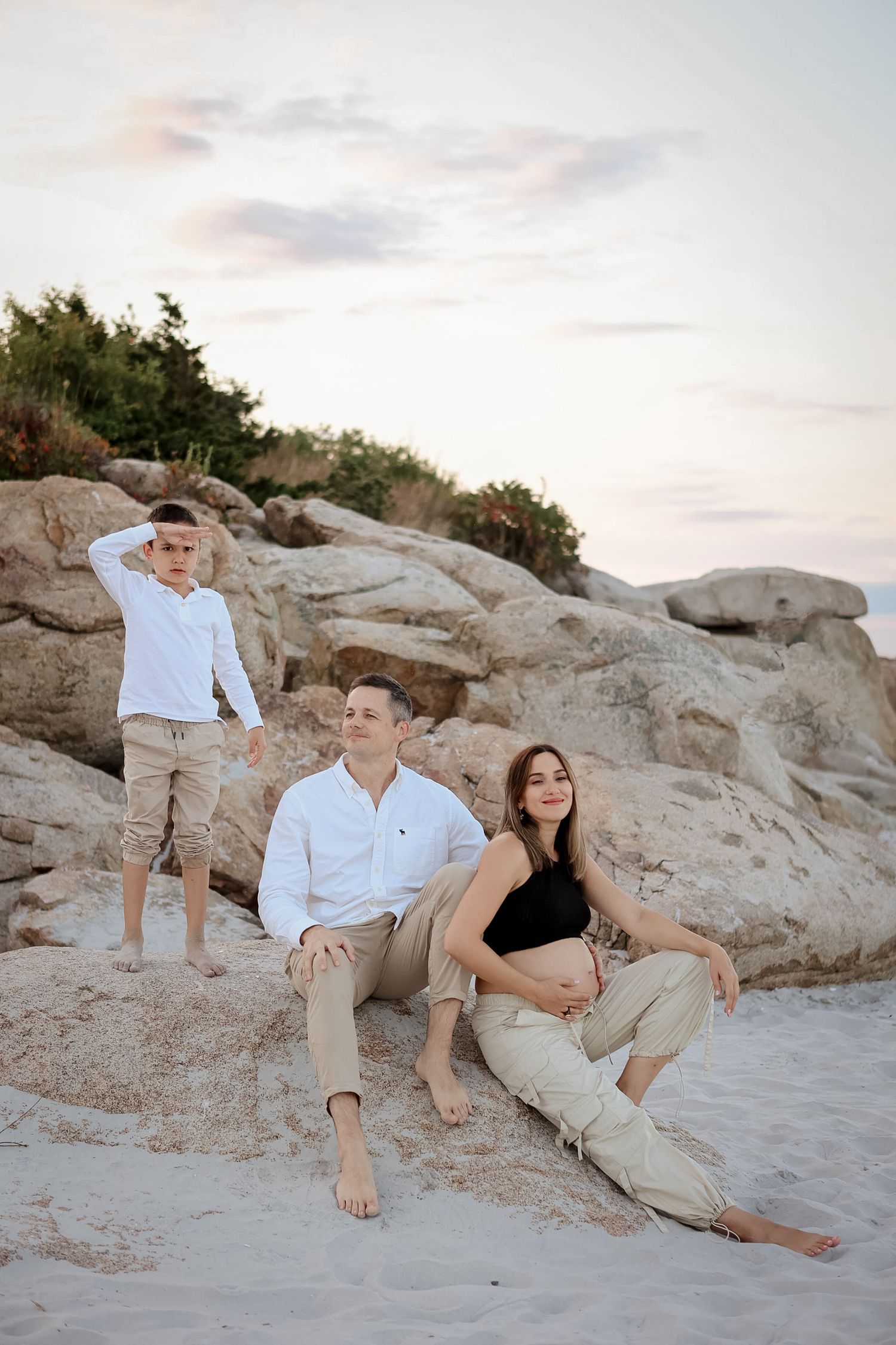 Pregnant woman with husband and son posing on rocky beach at sunset.