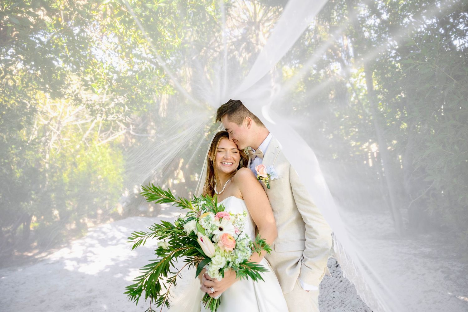 Bride and groom embrace under flowing veil in sunlit forest, bride holding lush white and pink bouquet.
