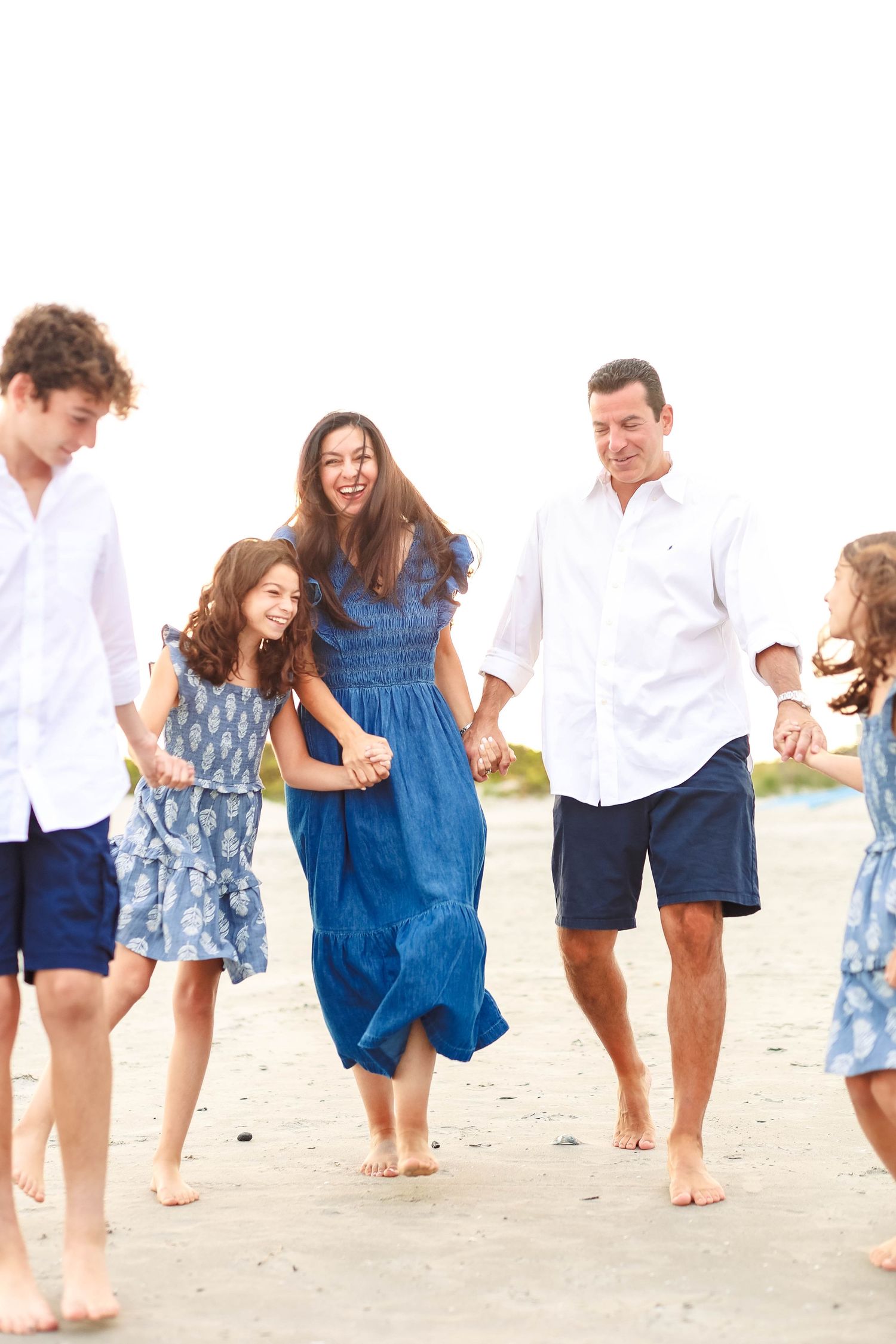 Happy family of five walking barefoot on a beach, dressed in coordinating blue and white outfits.