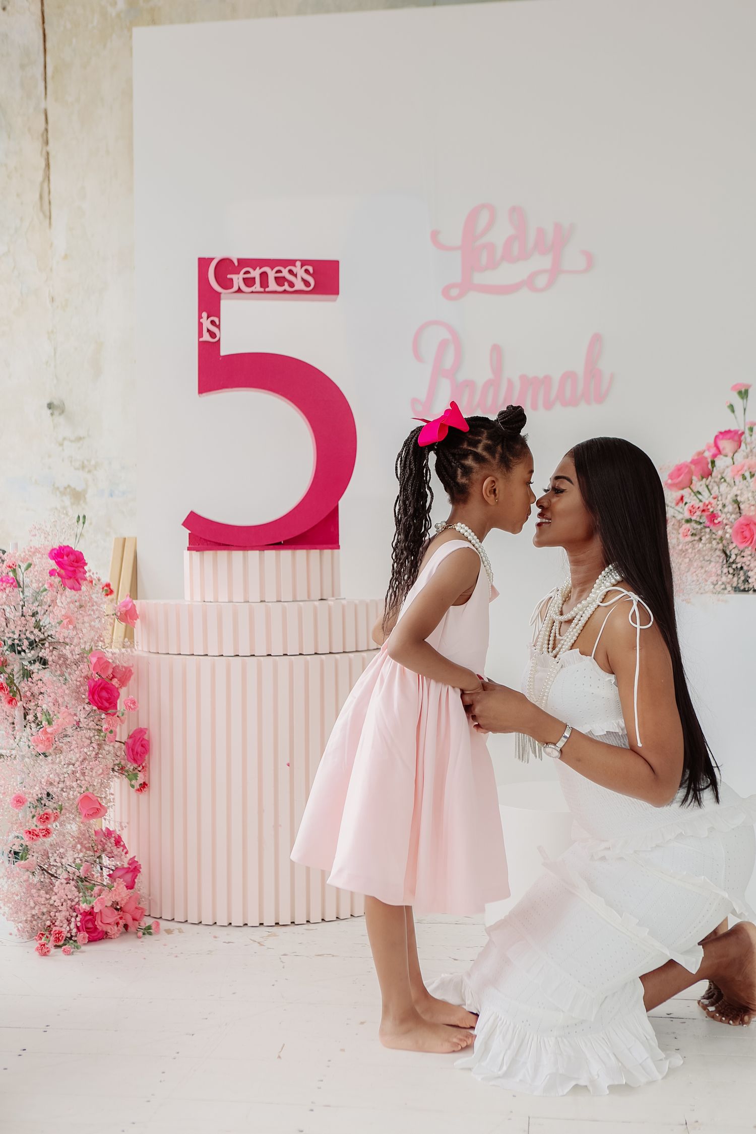 Mother in white dress kneeling to kiss daughter in pink skirt at Genesis Lady Padmah birthday party.