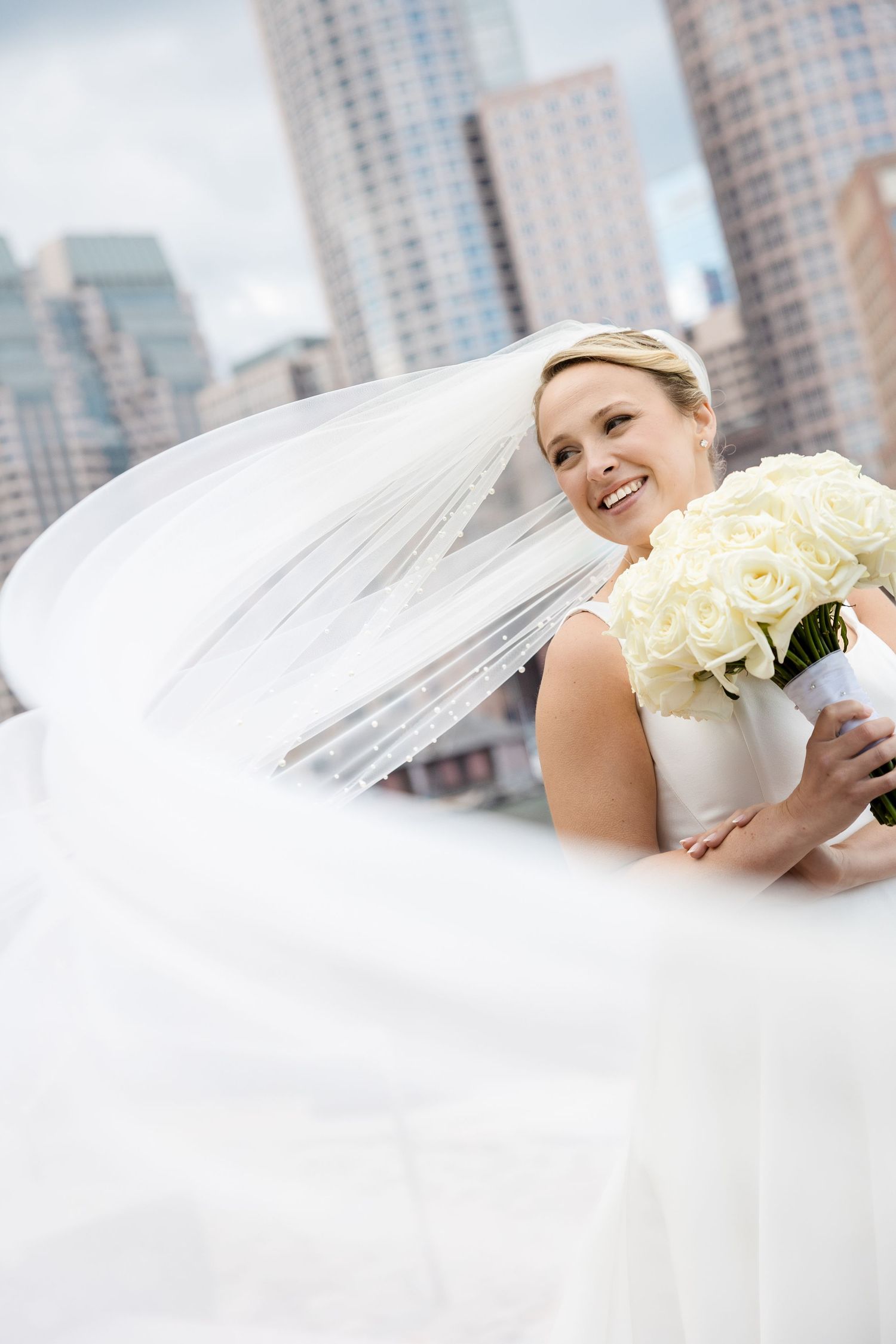 Bride in sleek white gown holding bouquet as veil flows at Boston Harbor Hotel in Boston, Massachusetts, captured by Grand Photo