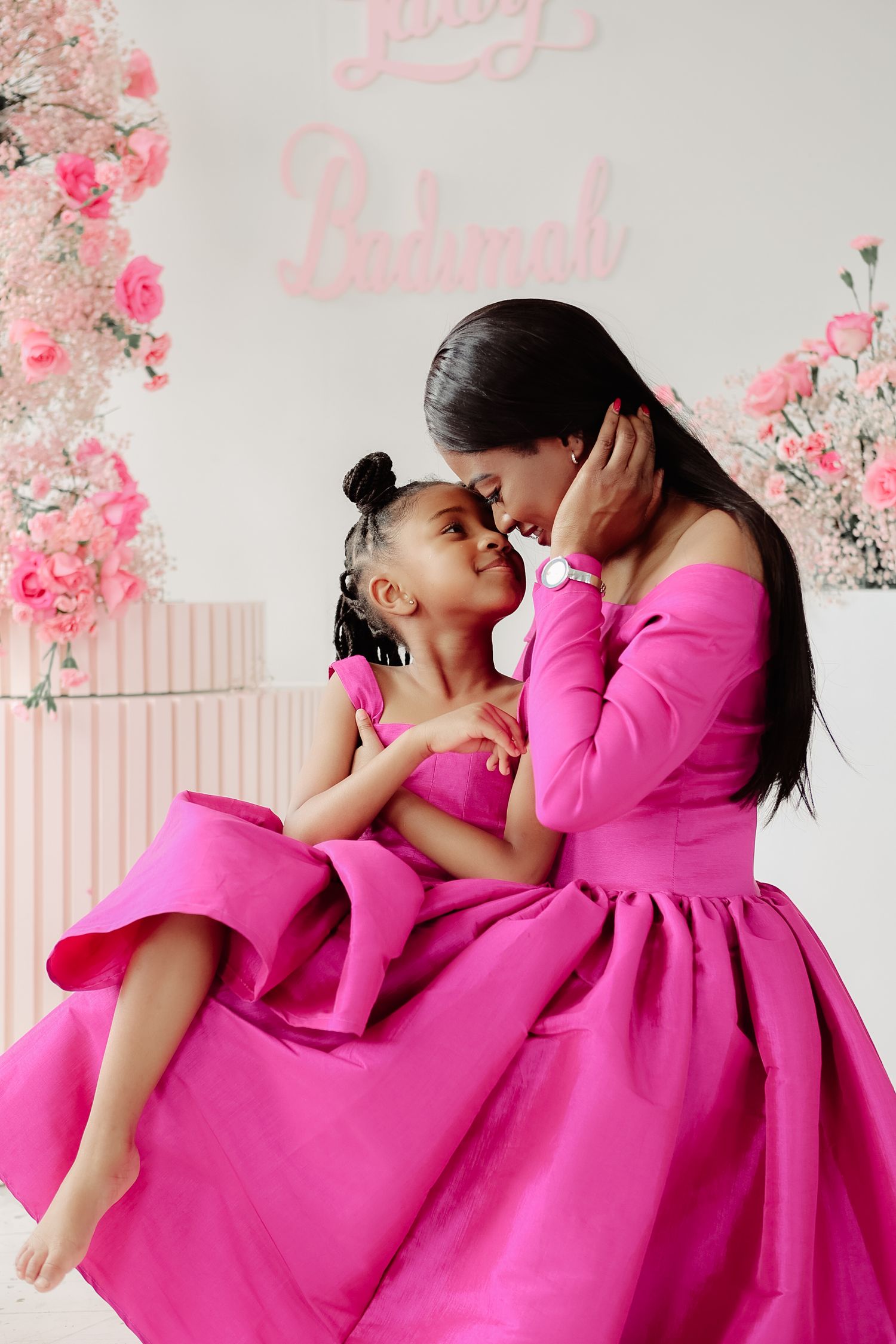 Mother and daughter in matching hot pink ball gowns embracing at floral birthday celebration backdrop.