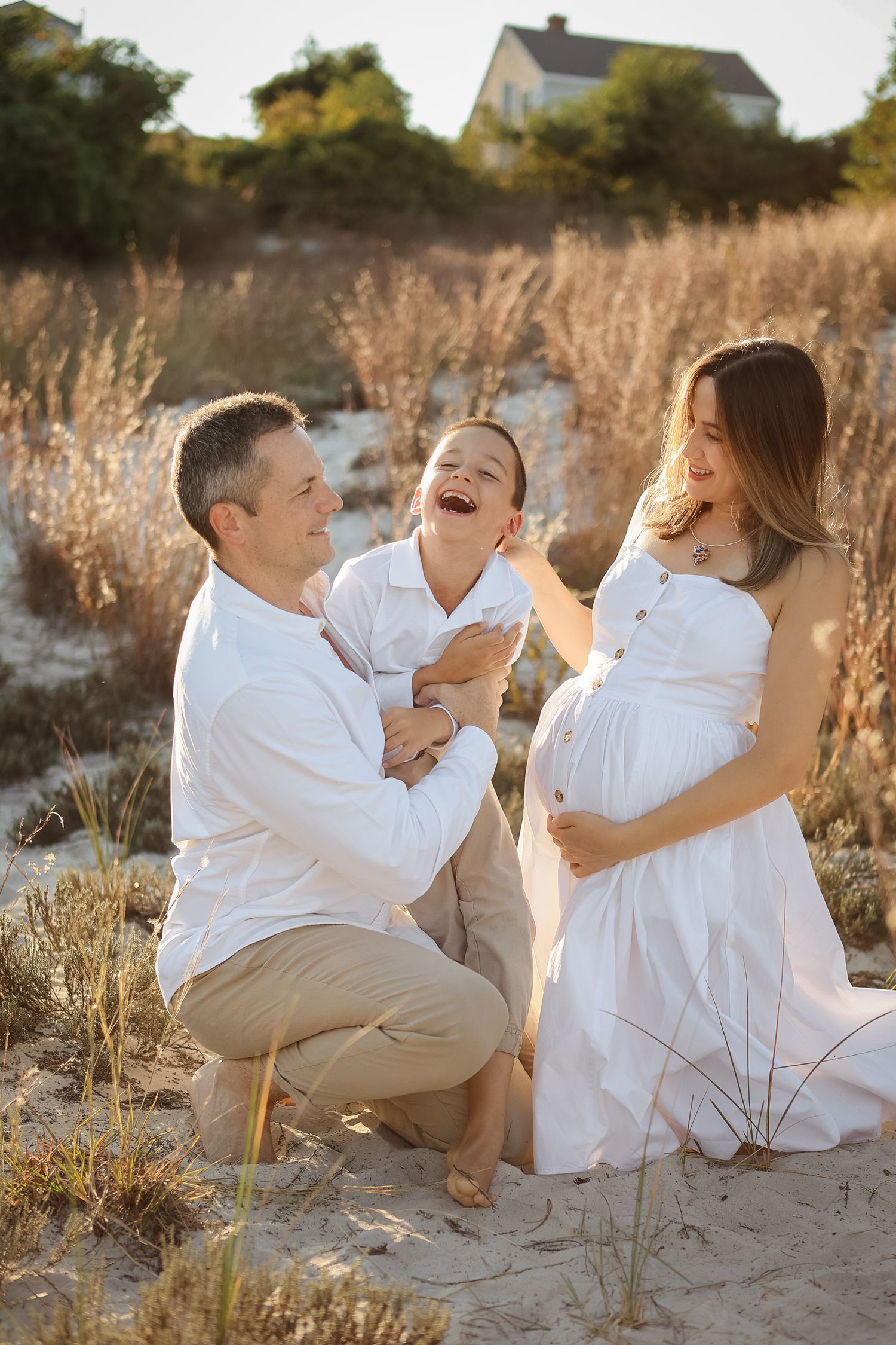 Happy family maternity photo on sandy beach at golden hour with pregnant mother in white dress.