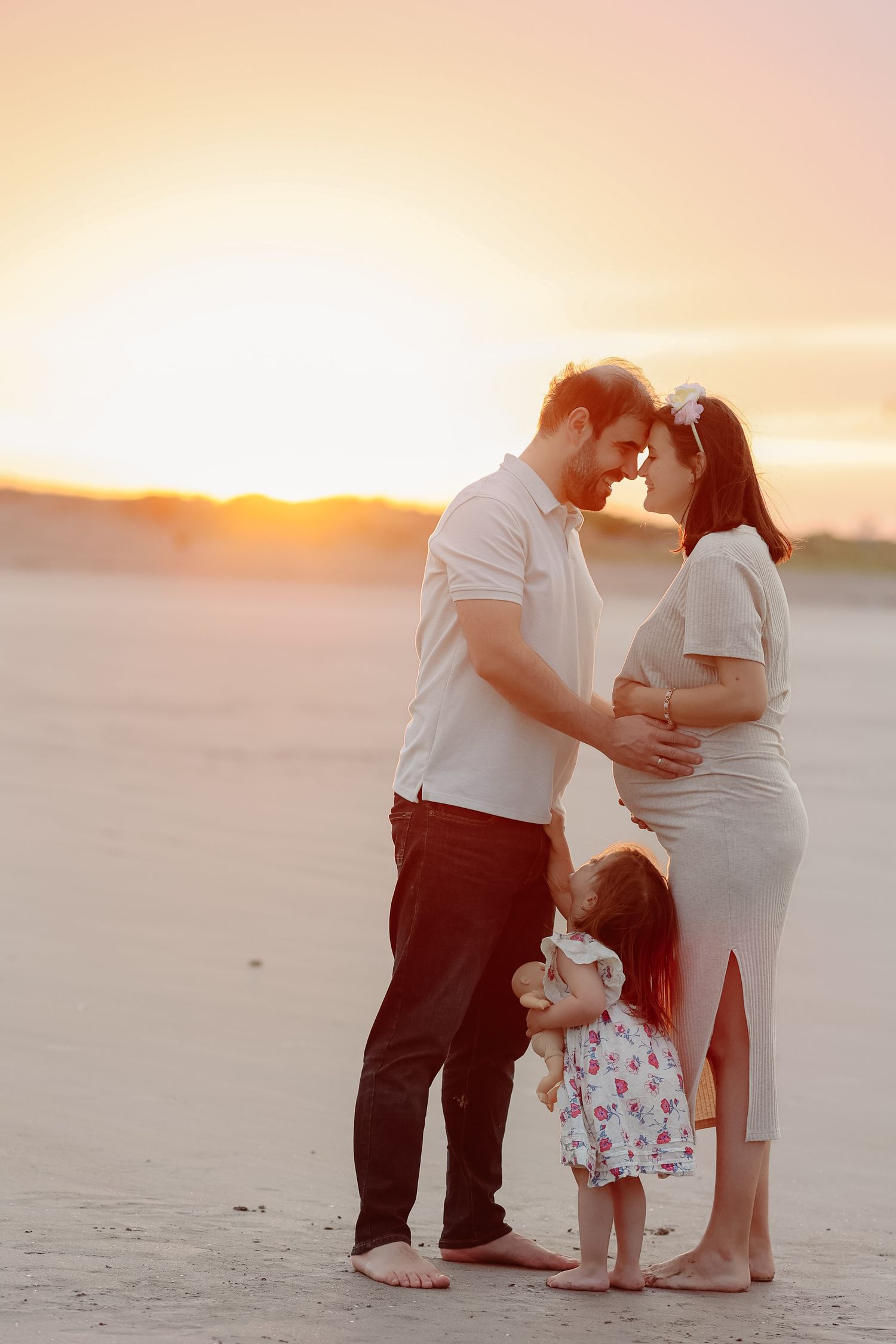 Family maternity photo at sunset on beach, couple touching foreheads with toddler daughter standing between them.
