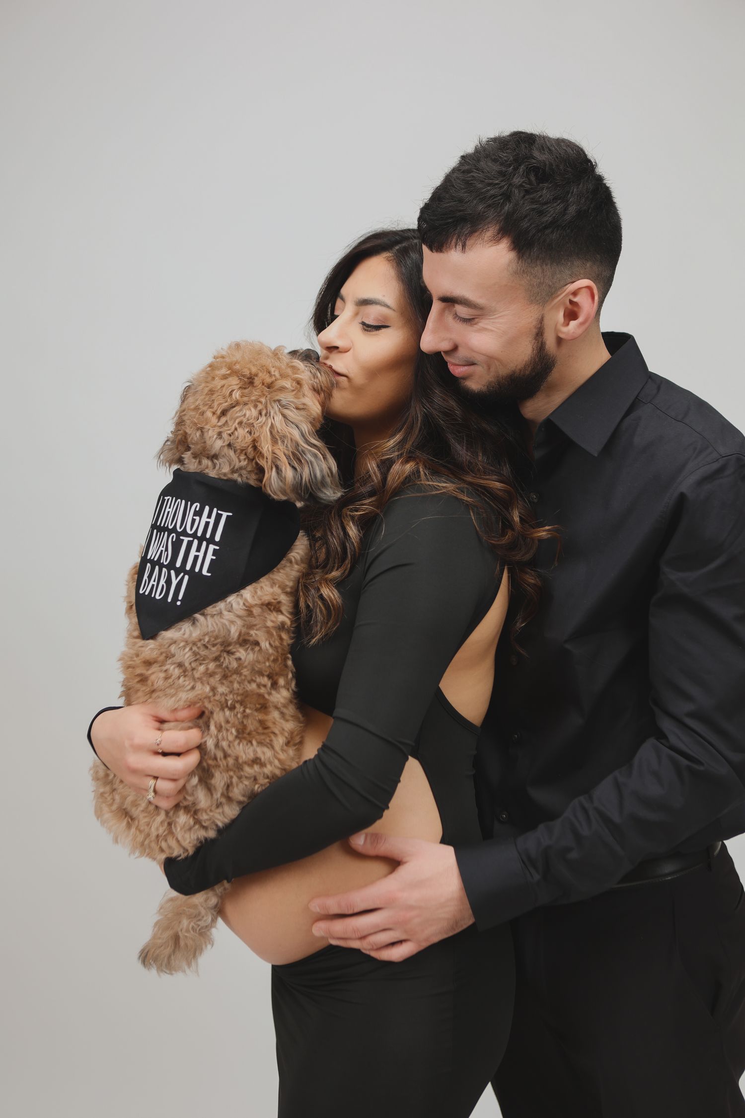 Pregnant couple in black outfits kissing golden doodle dog with baby bandana in studio.