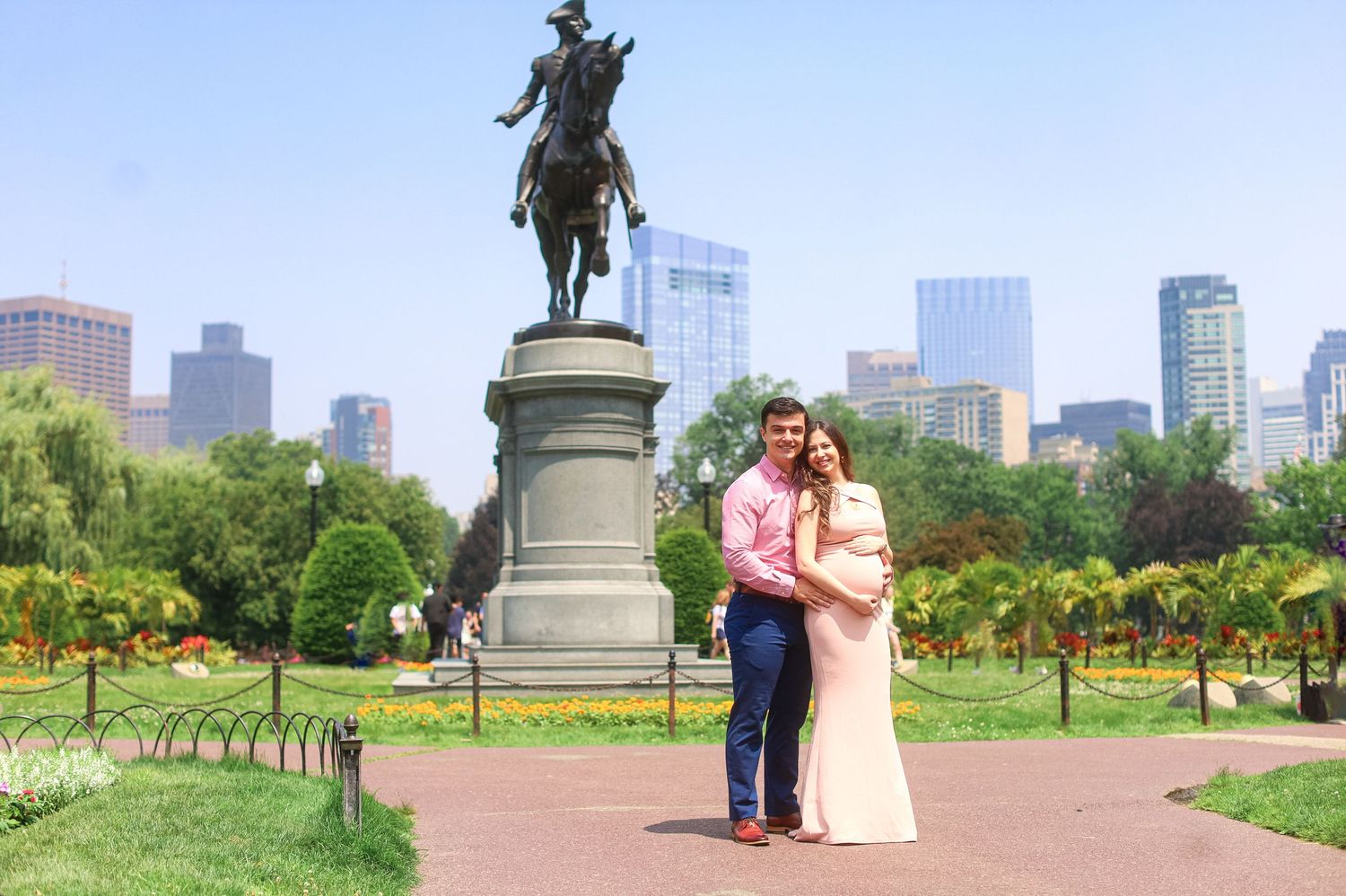 Smiling pregnant couple poses in front of George Washington statue in Boston Public Garden on sunny day.