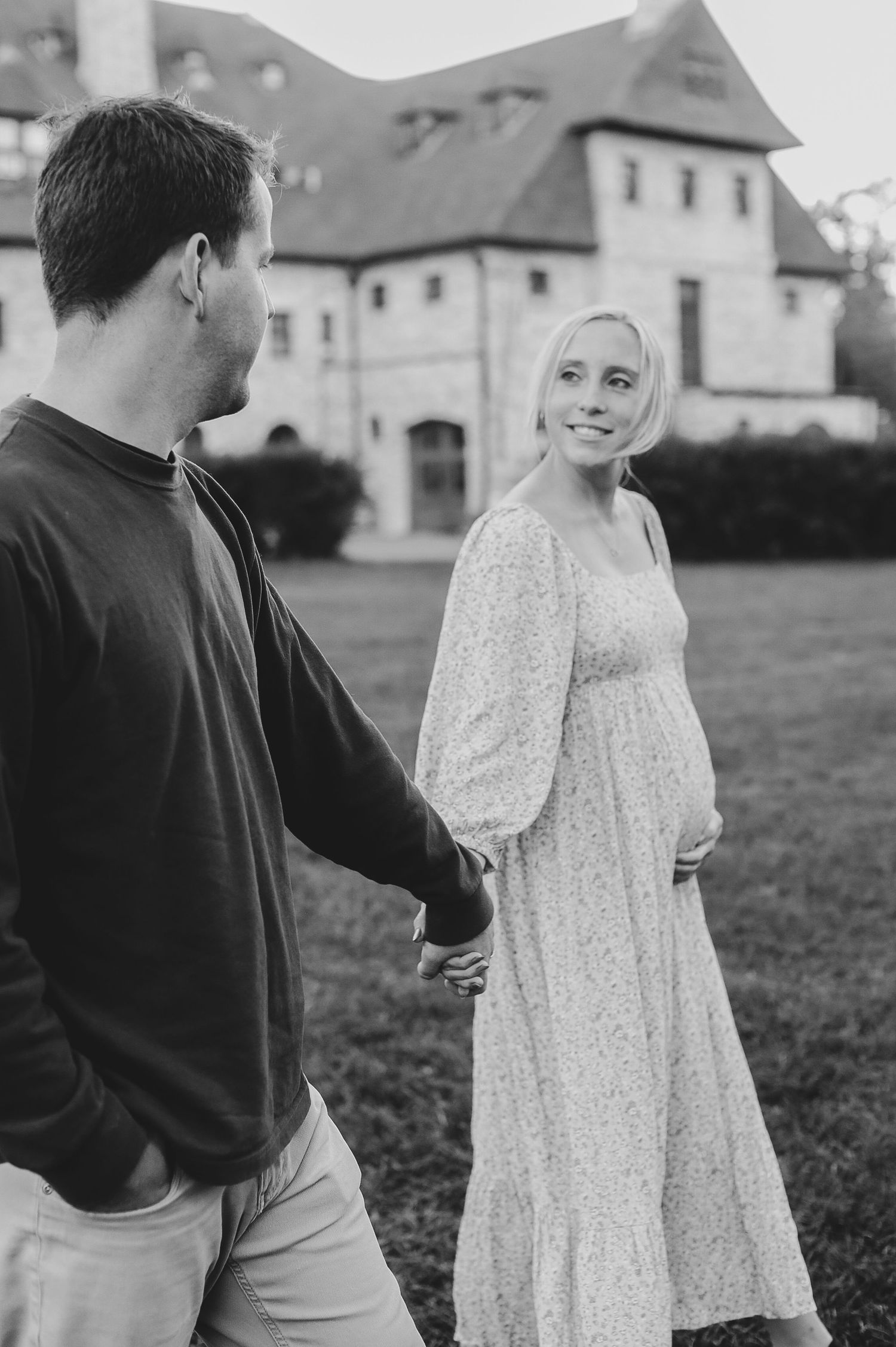Black and white photo of pregnant couple holding hands walking near historic building at dusk.