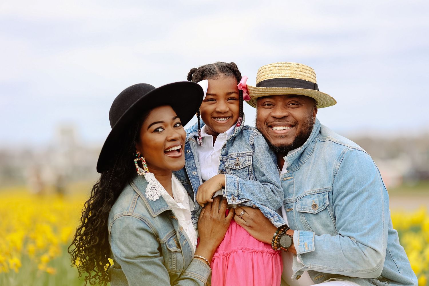 Happy Black family smiling outdoors in yellow flower field, parents holding young daughter in pink dress.