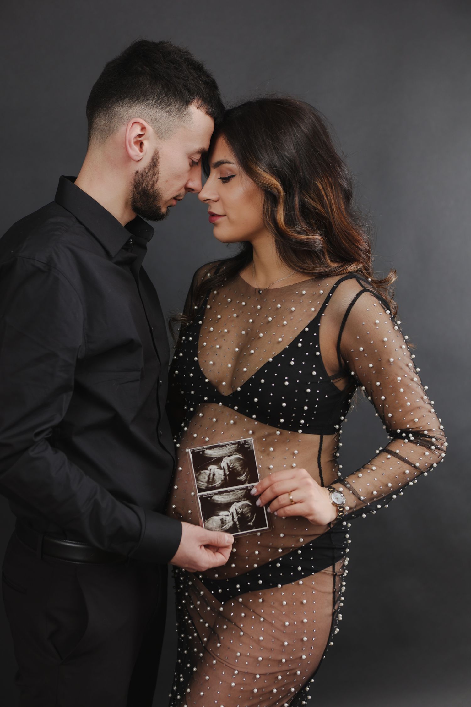 Couple in black outfits sharing an intimate moment while holding ultrasound photos during maternity shoot.