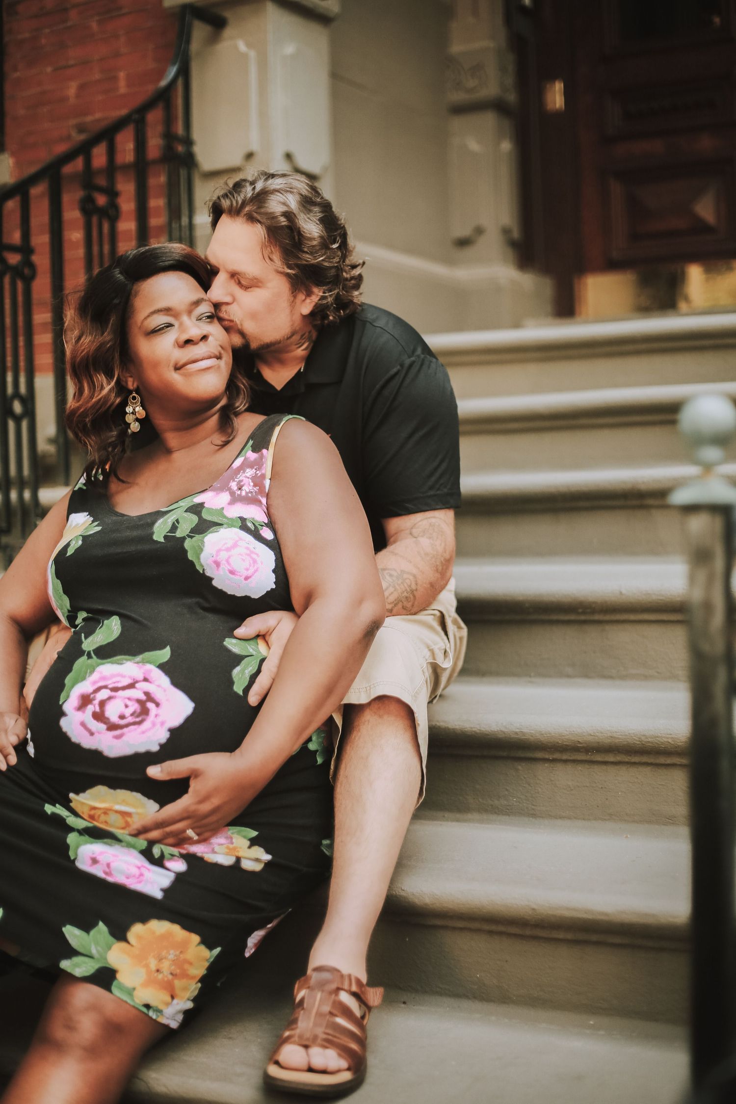 Interracial couple sitting on urban stoop steps, man kissing pregnant woman in floral dress.