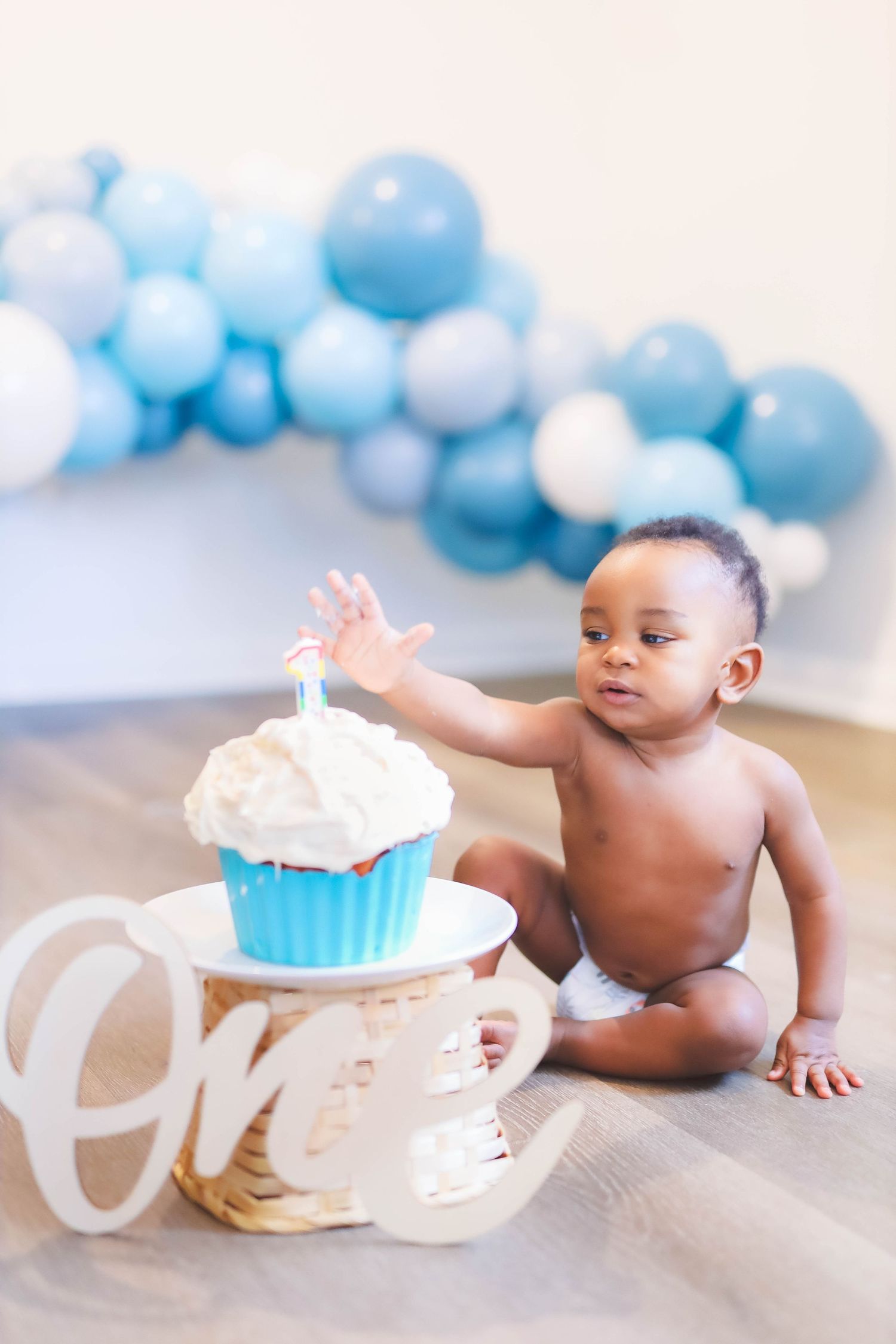 Baby touching first birthday cupcake during cake smash with blue balloon garland backdrop.