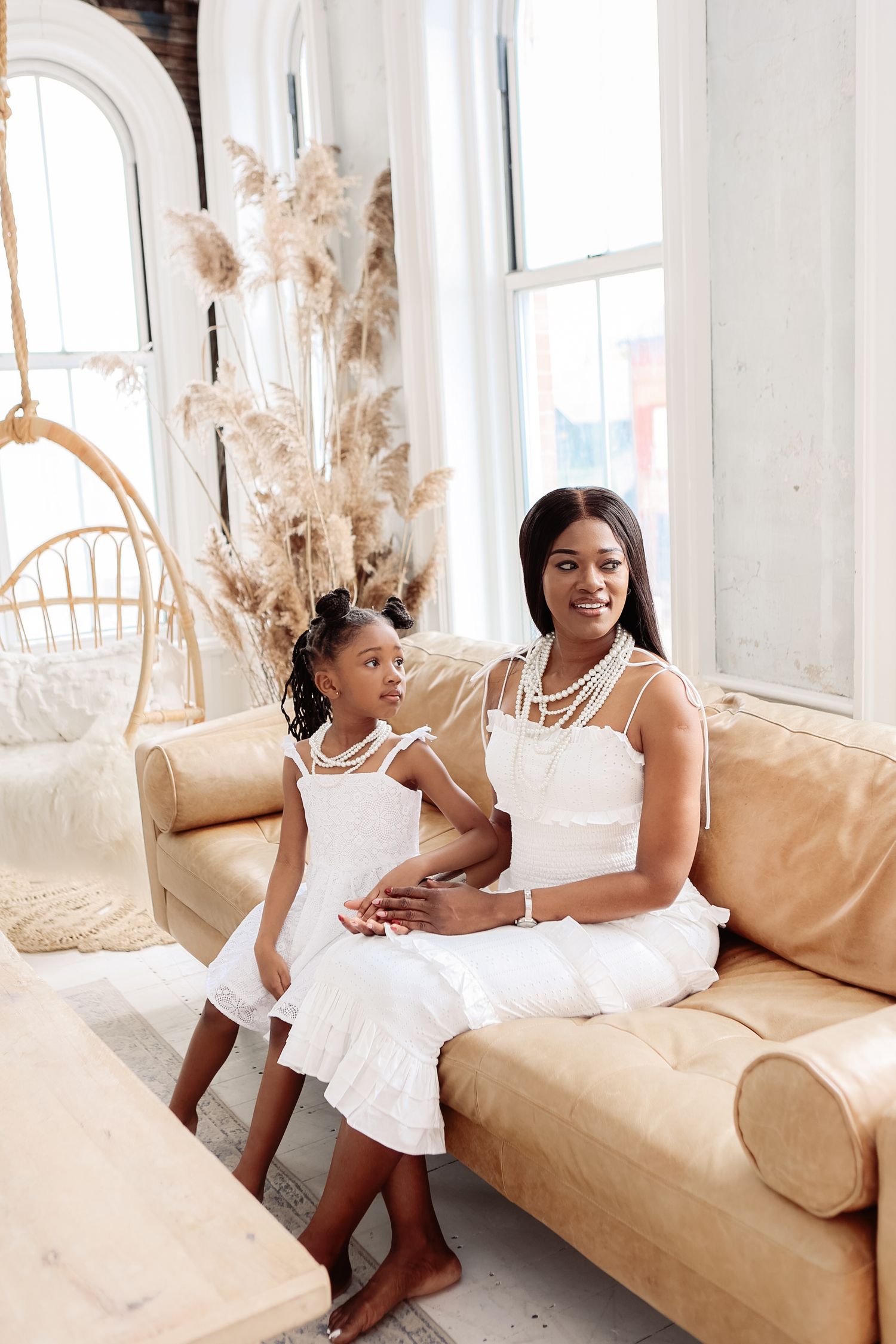 Mother and daughter in matching white ruffle dresses with pearls sitting on tan leather sofa.