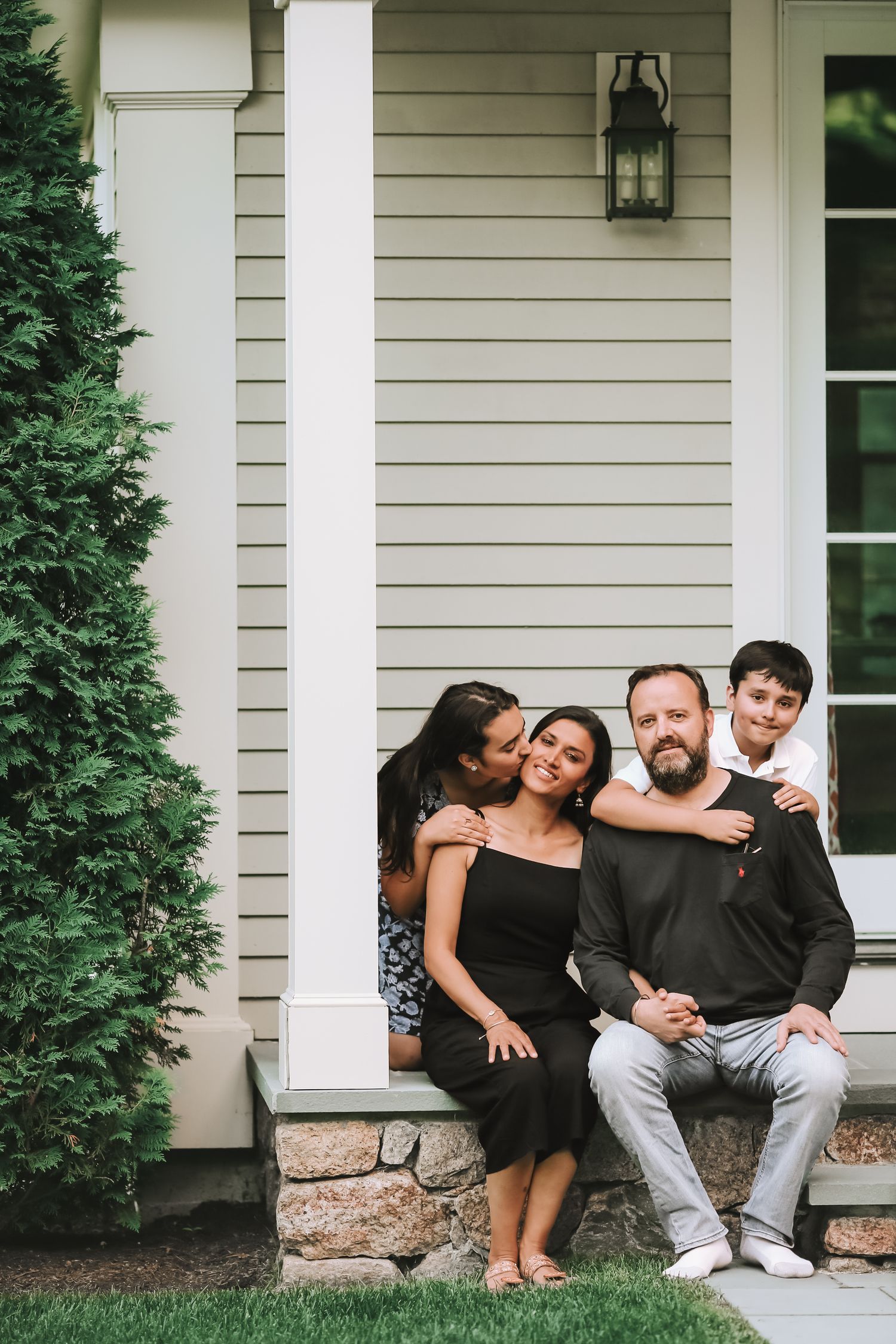 Happy family of four posing on front porch steps of a classic white suburban home.