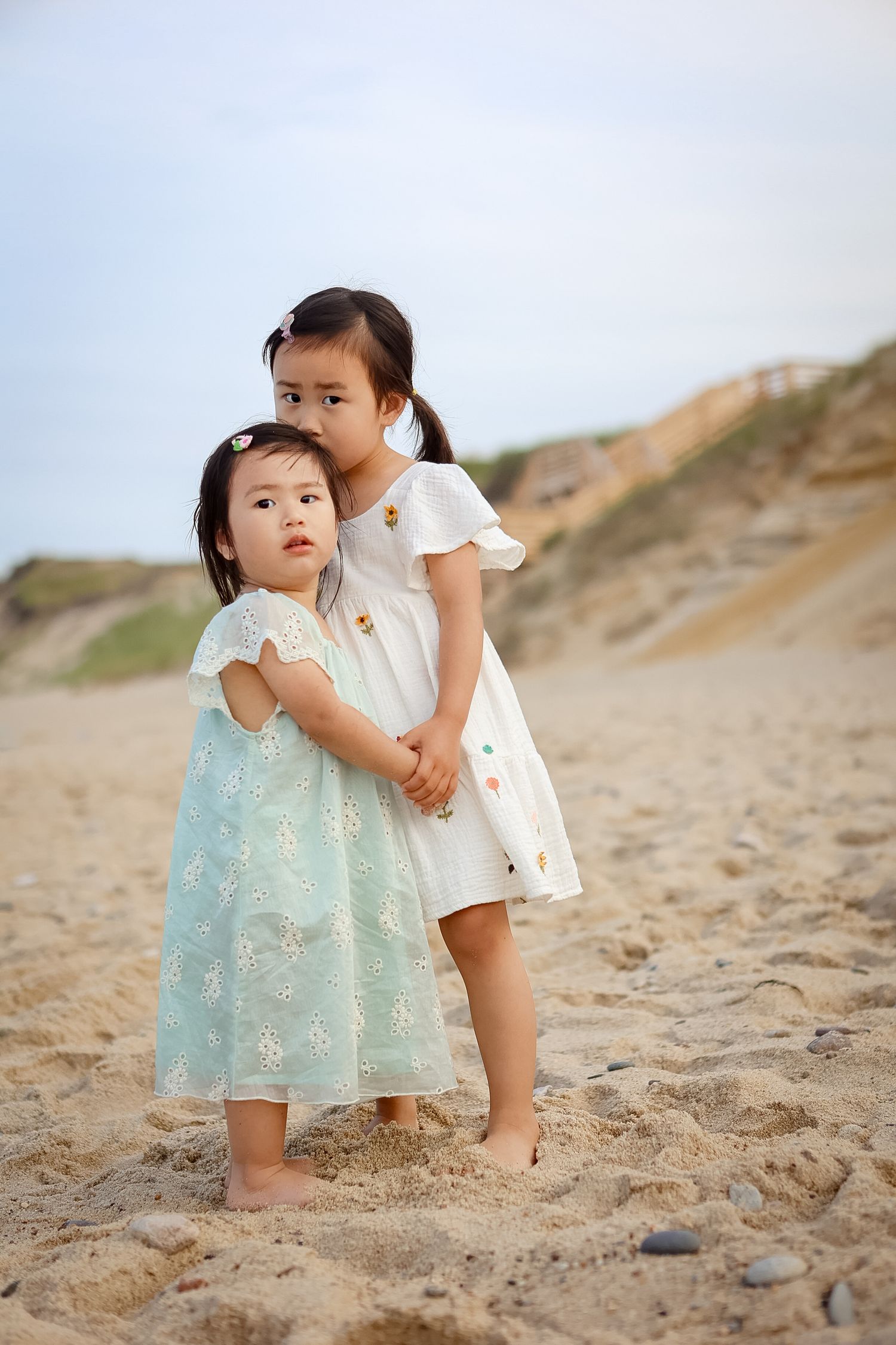 Two young Asian sisters in white and teal dresses standing together on sandy beach.