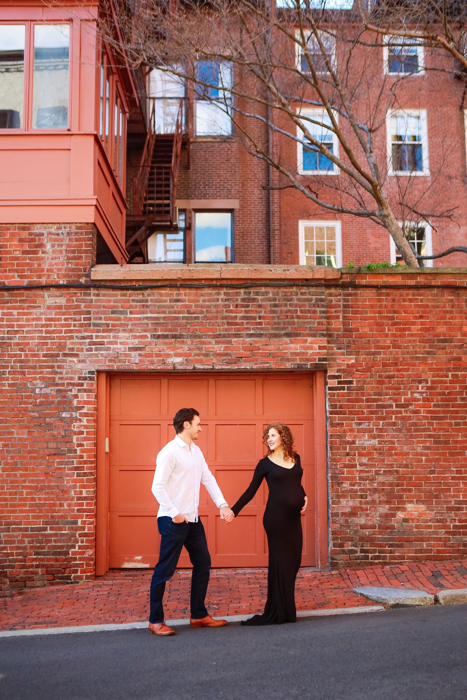 Expecting couple holds hands in front of red brick building and garage door during outdoor maternity photoshoot.