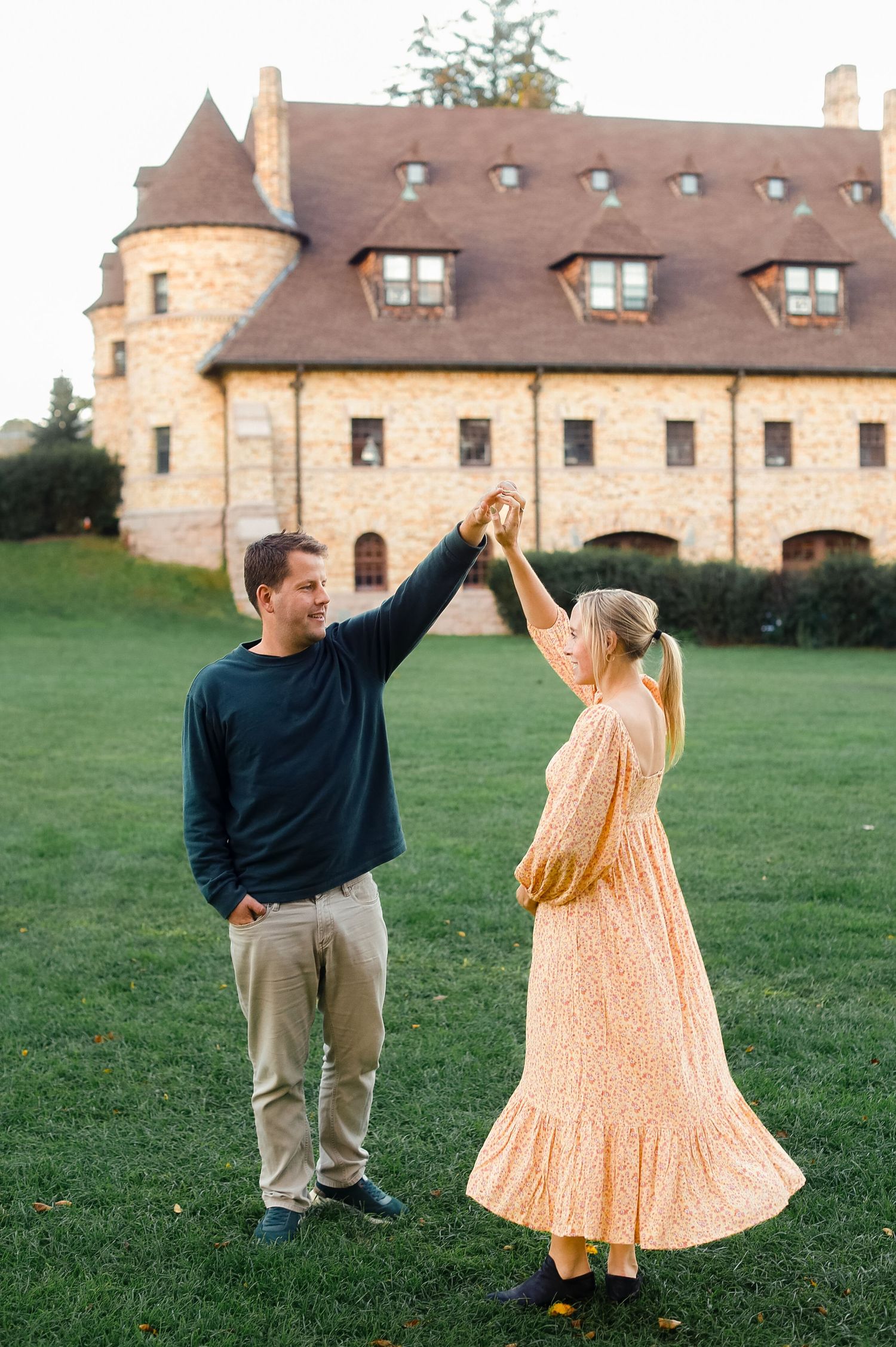 Happy couple dances on green lawn in front of charming stone castle at golden hour.