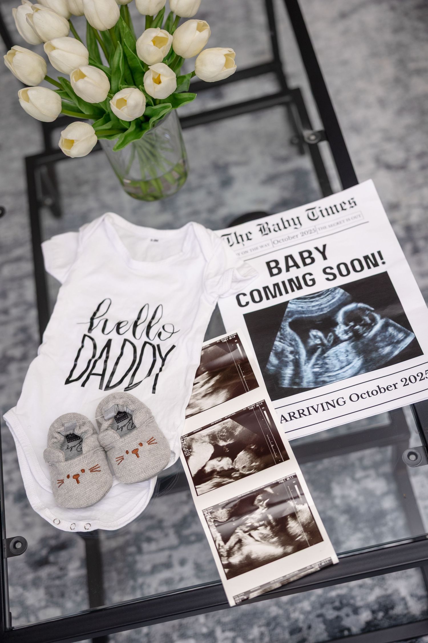 Baby announcement flat lay with Hello Daddy onesie, ultrasound photos, booties, and white tulips.
