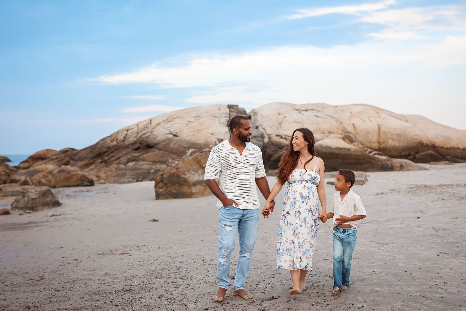 Interracial family of three walks hand in hand on sandy beach with rocky cliffs behind.