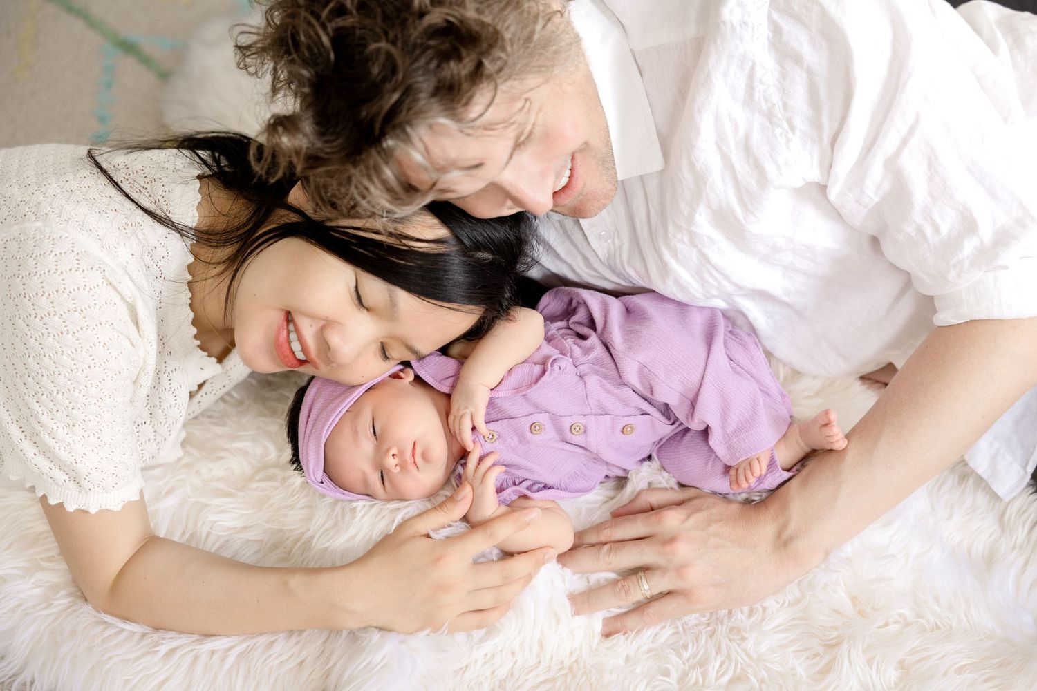 Parents kissing newborn baby wrapped in purple swaddle on white bed.