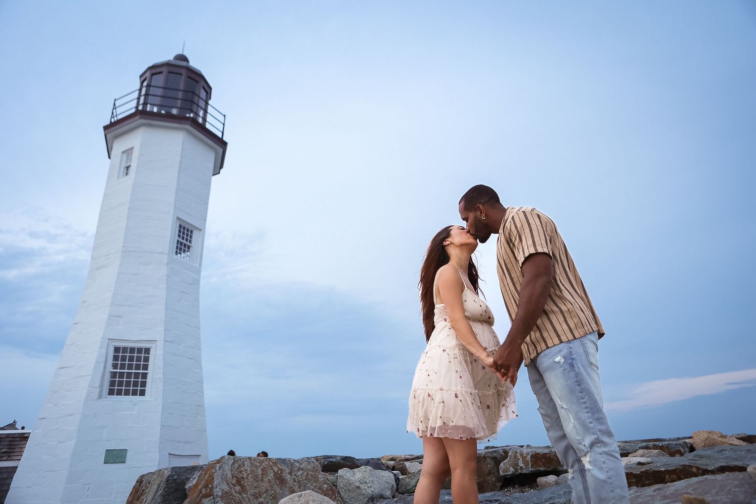 Interracial couple sharing a tender moment near a lighthouse at dusk on rocky shore.