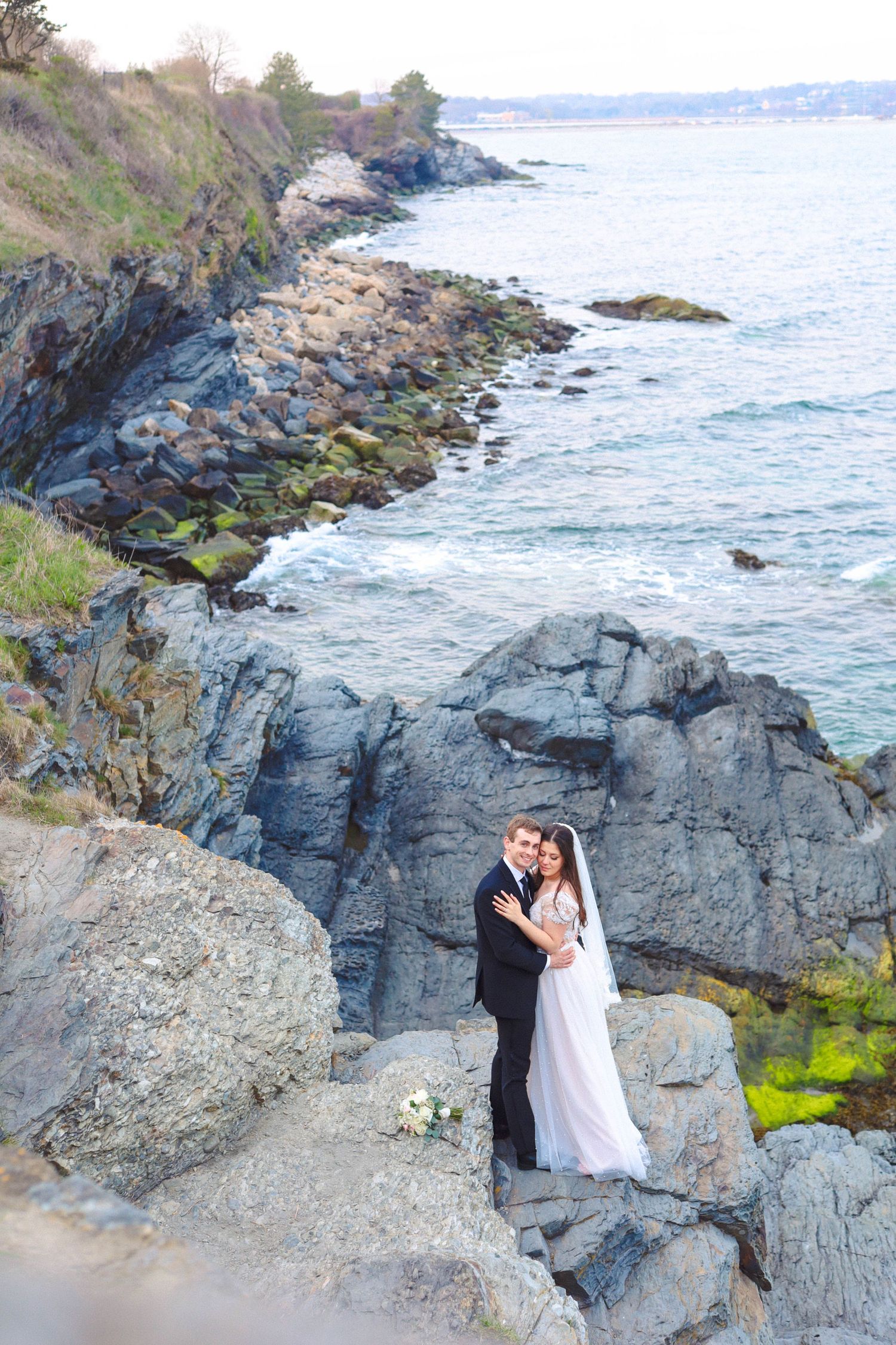 Wedding couple embracing on rocky coastal cliffs at The Chanler at Cliff Walk in Newport, Rhode Island, captured by Grand Photo