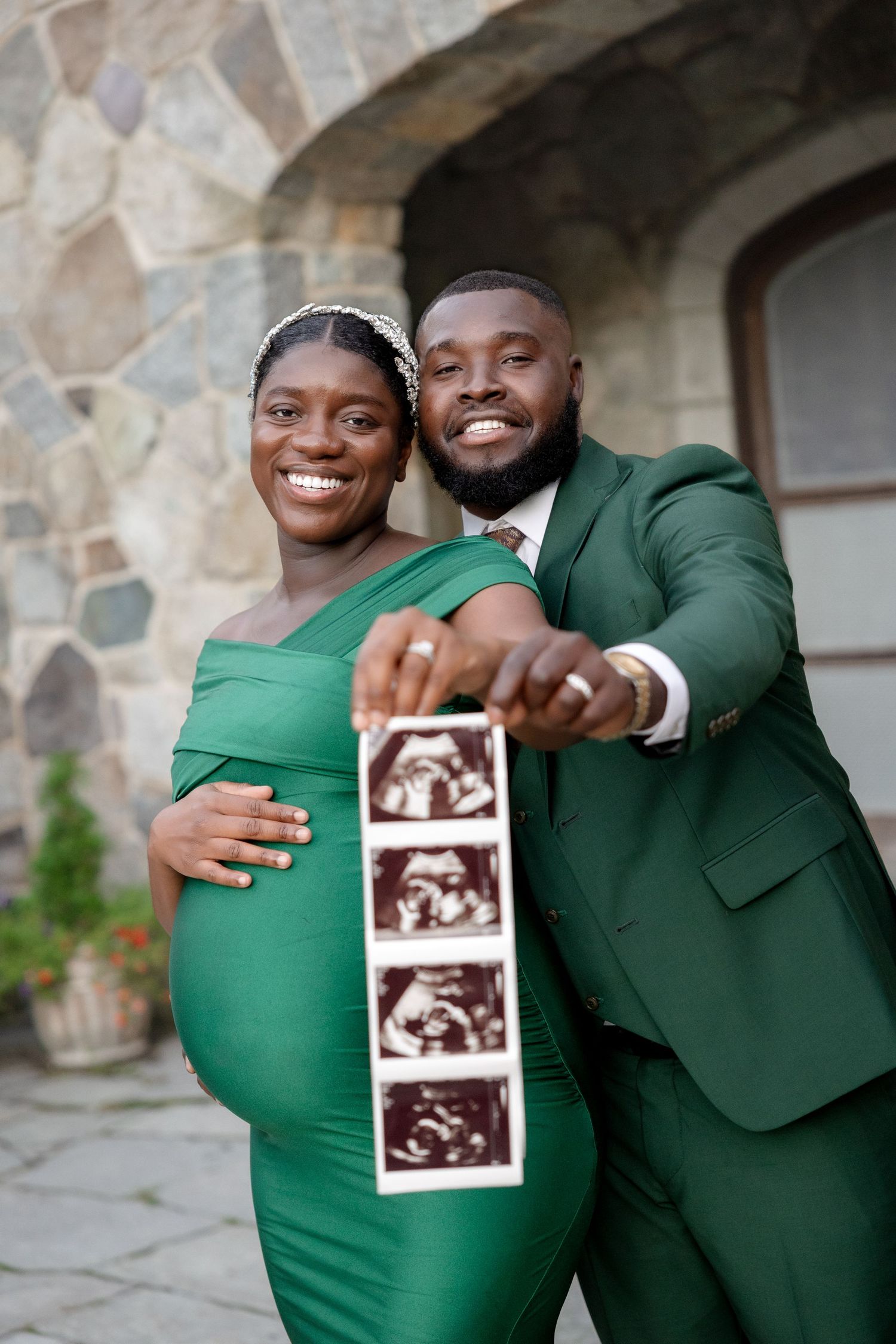 Smiling couple in green outfits proudly displaying ultrasound images during outdoor maternity photoshoot.
