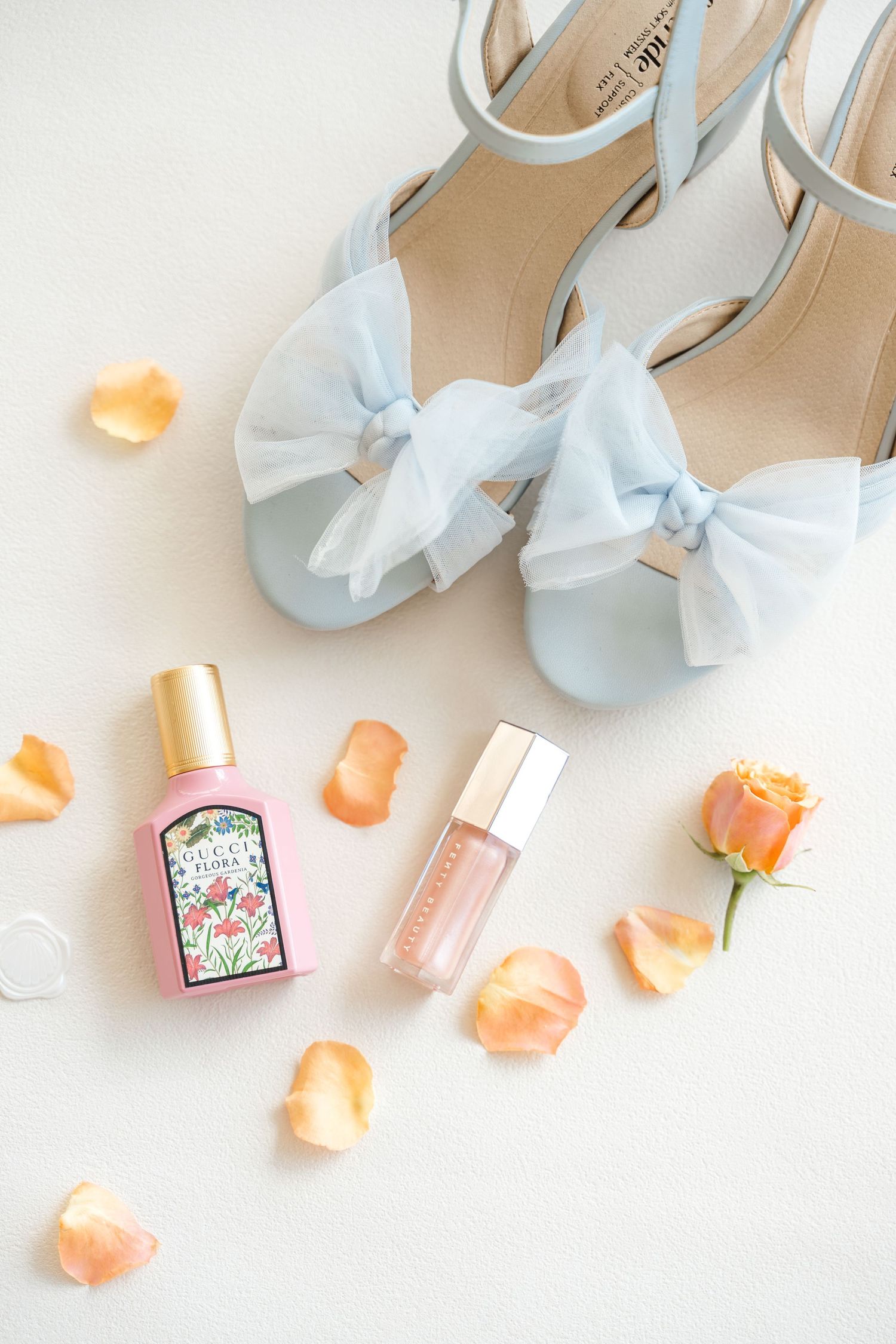 Light blue bow heeled sandals with Gucci Flora perfume, pink lip gloss, and orange rose petals on white background.