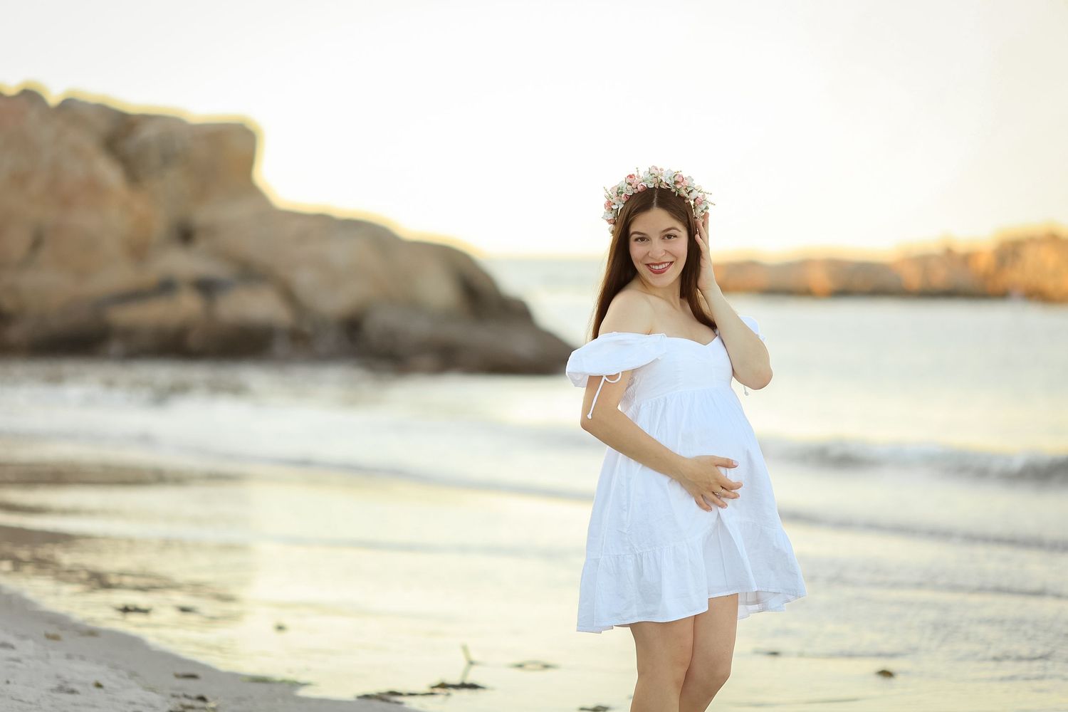 Smiling pregnant woman in white off-shoulder dress and floral crown walks along a sunny beach shoreline.