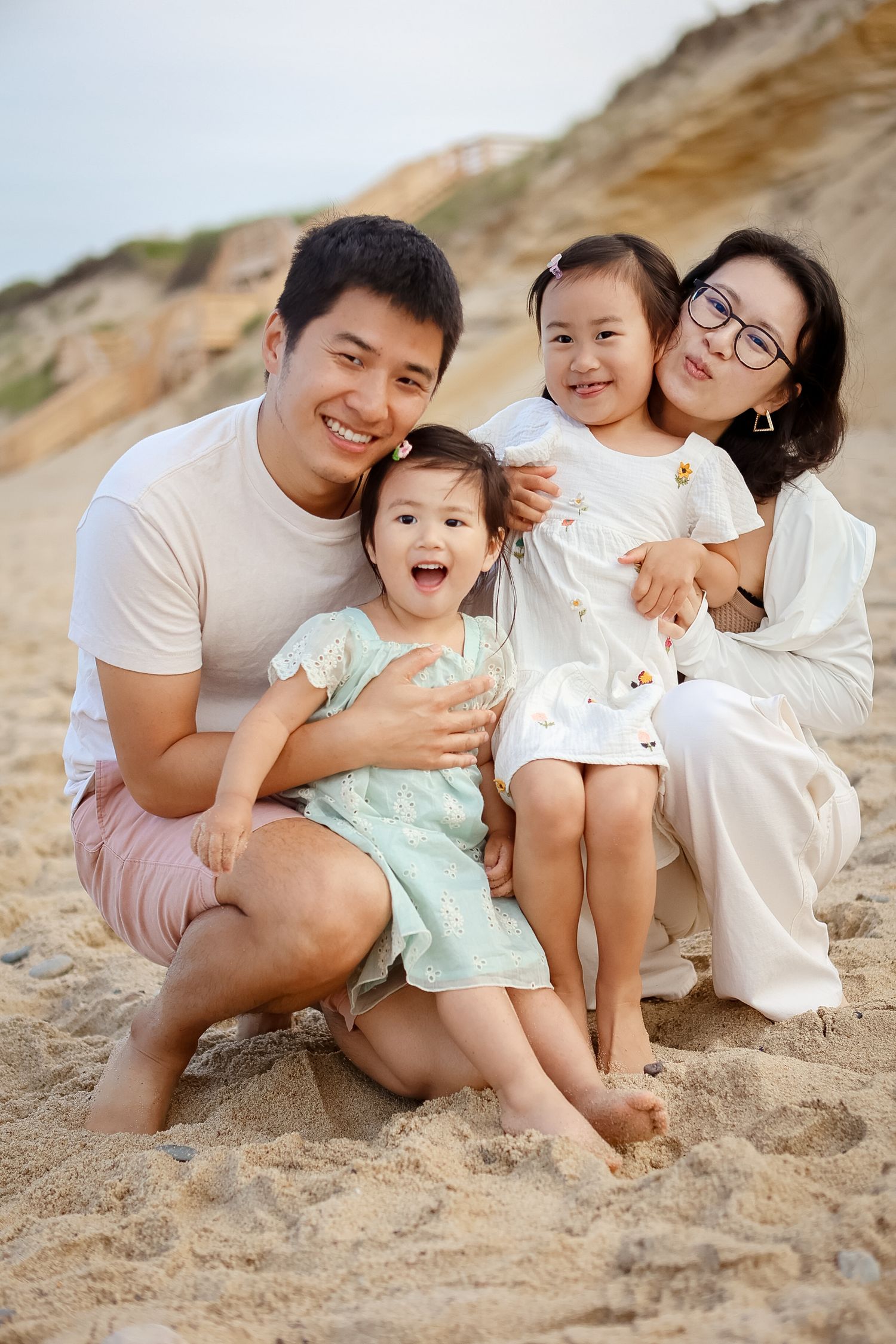 Happy Asian family of four smiles together sitting on sandy beach with sand dunes behind.