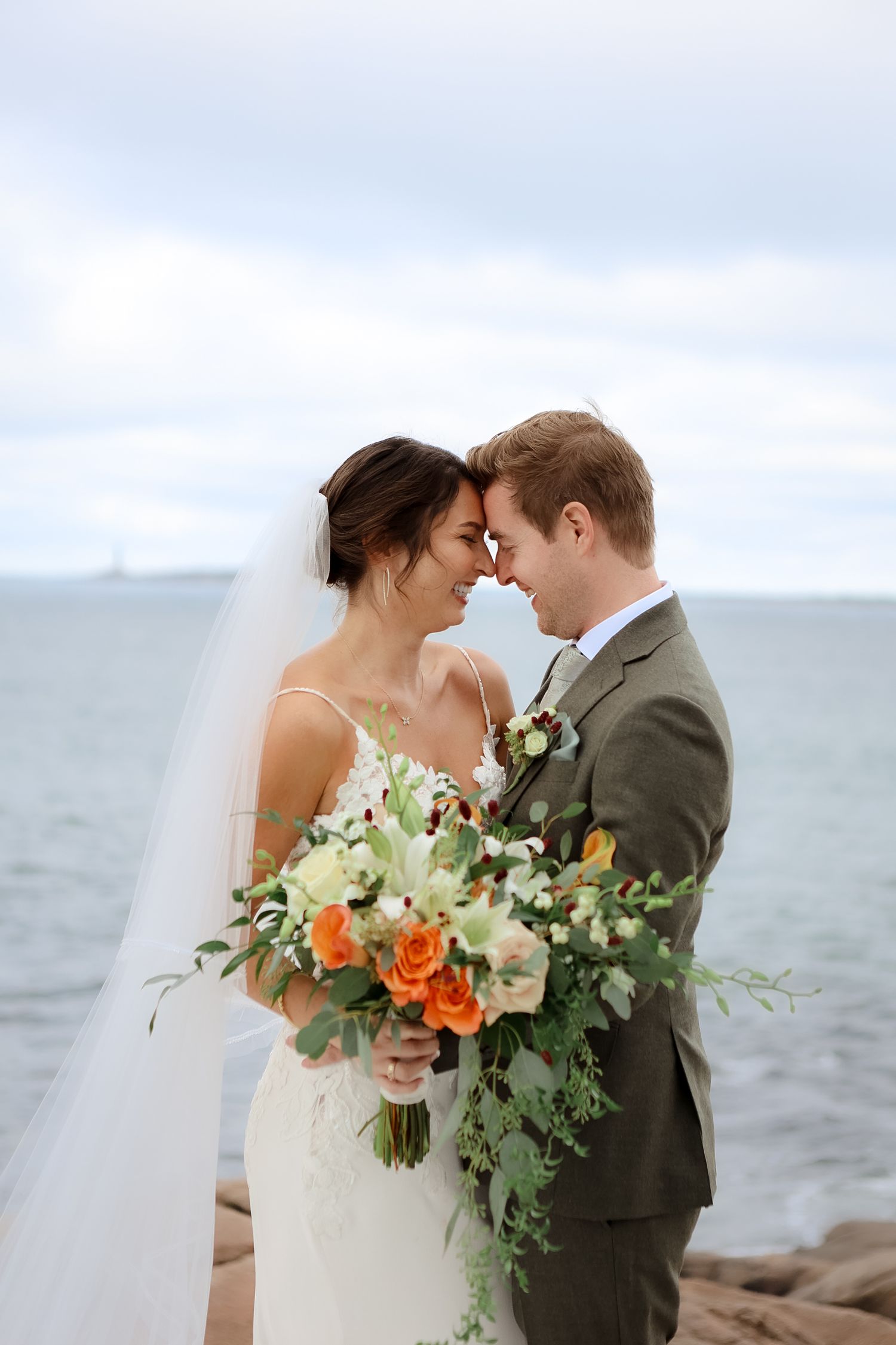 Bride and groom touch foreheads romantically holding orange floral bouquet with ocean backdrop on wedding day.