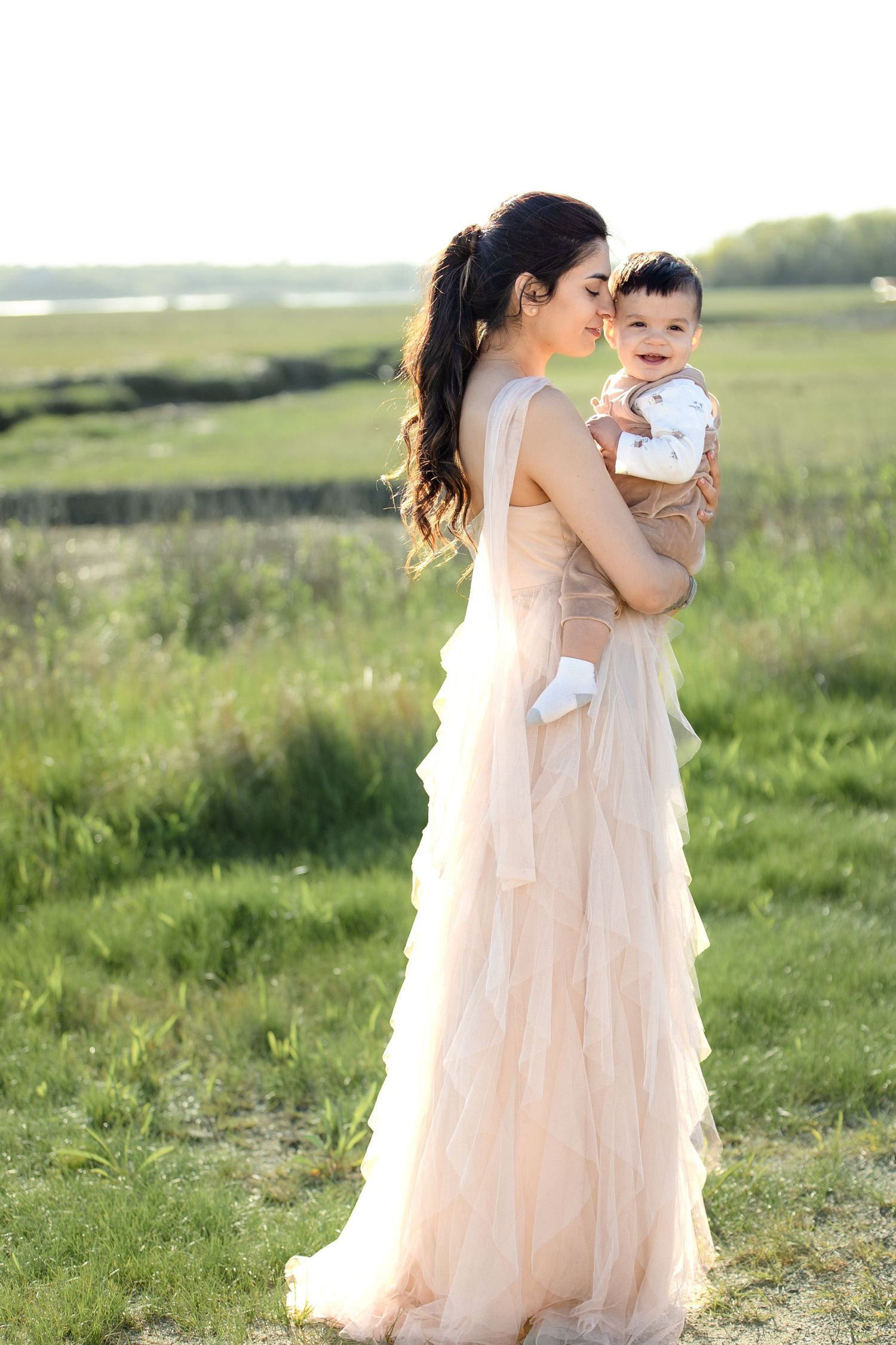 Elegant mother in pink gown holding baby in sunny green field.