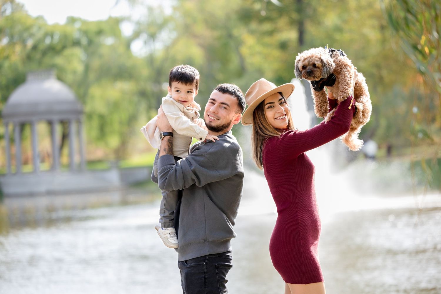 Couple holding baby and small dog by fountain in fall park.