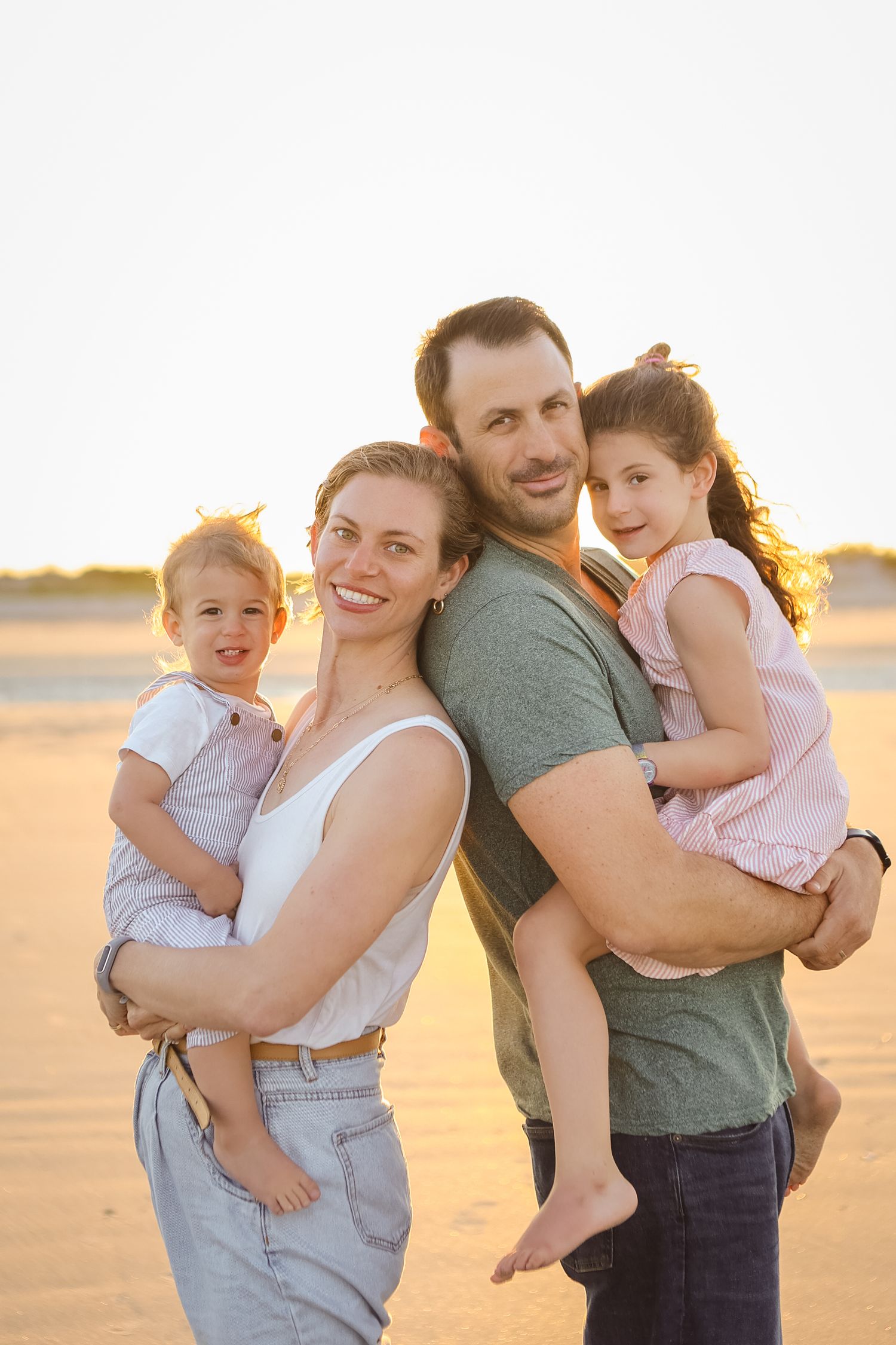Smiling family of four posing on sandy beach at golden sunset, parents holding two young daughters.