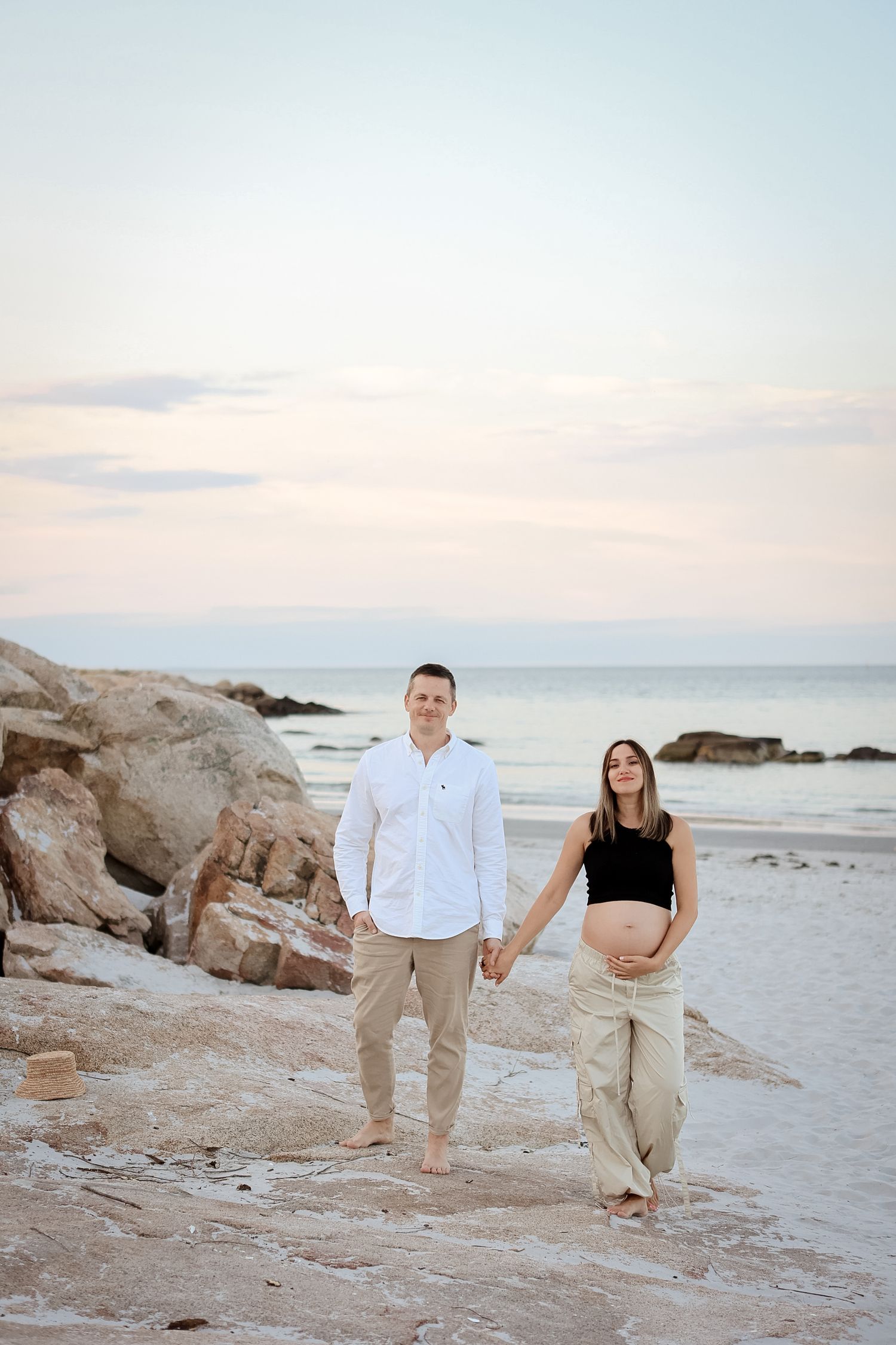 Couple holding hands on rocky beach at sunset, pregnant woman in black top and tan pants.