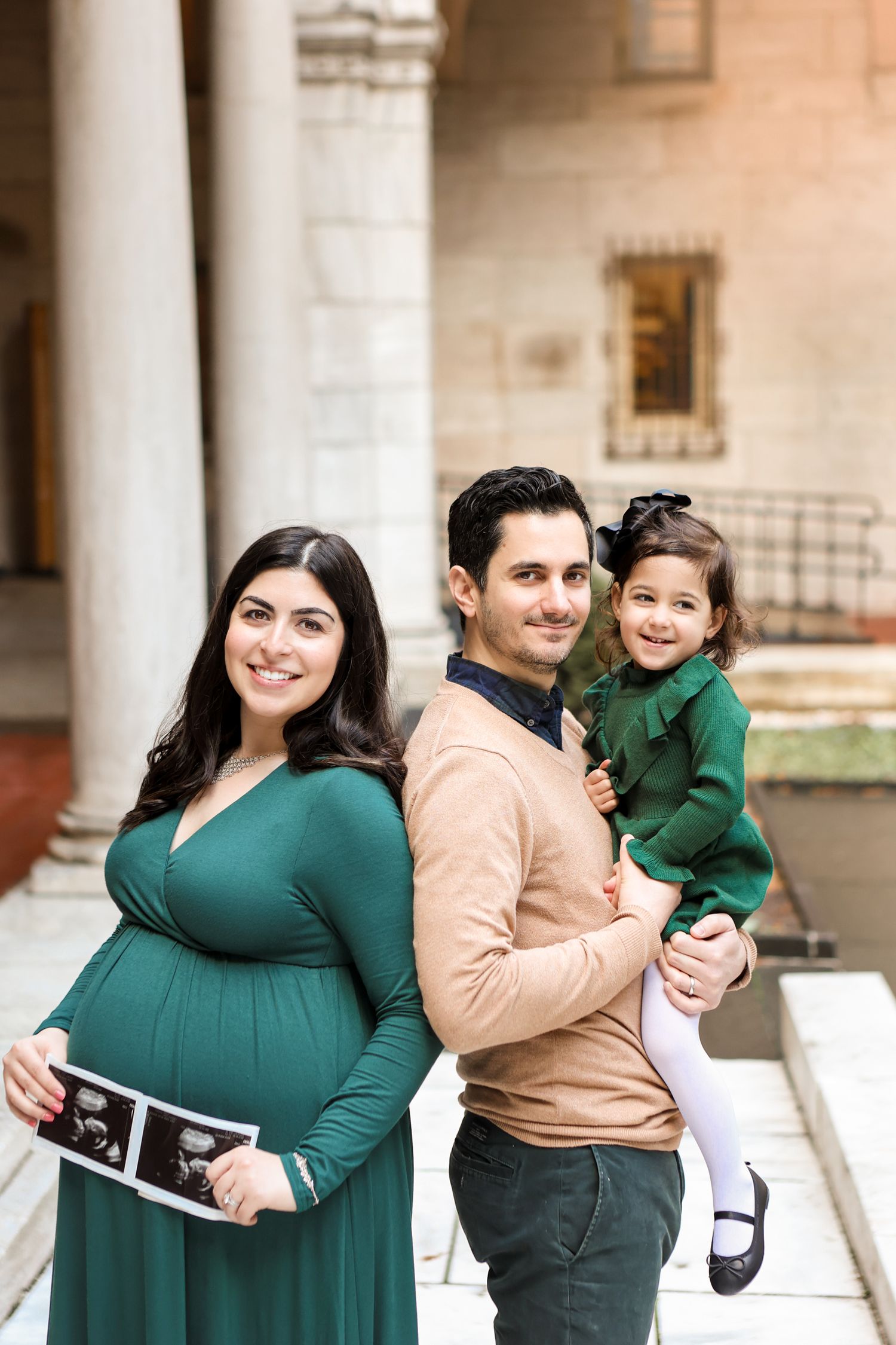 Pregnant woman in green dress holds ultrasound photo with husband and toddler daughter during family maternity photoshoot.