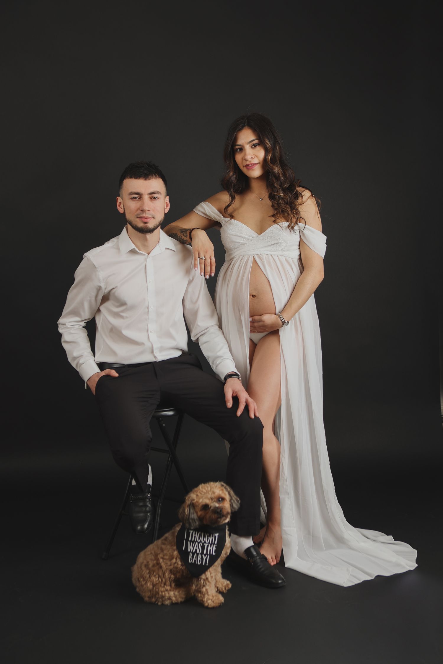 Expecting couple in white and black outfits posing with golden doodle dog in studio.