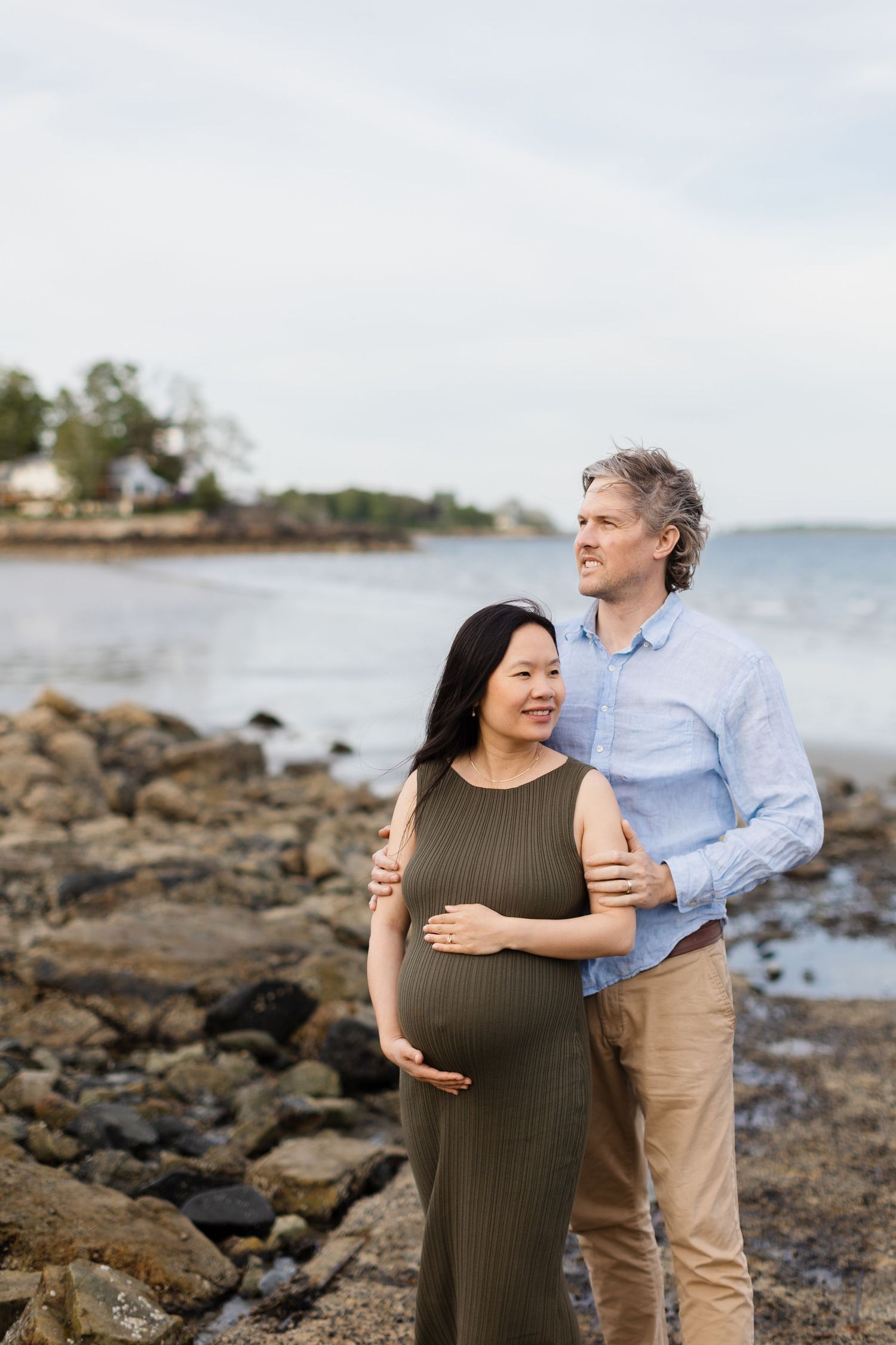 Pregnant woman in olive dress and partner embrace while gazing at ocean during coastal maternity photoshoot.
