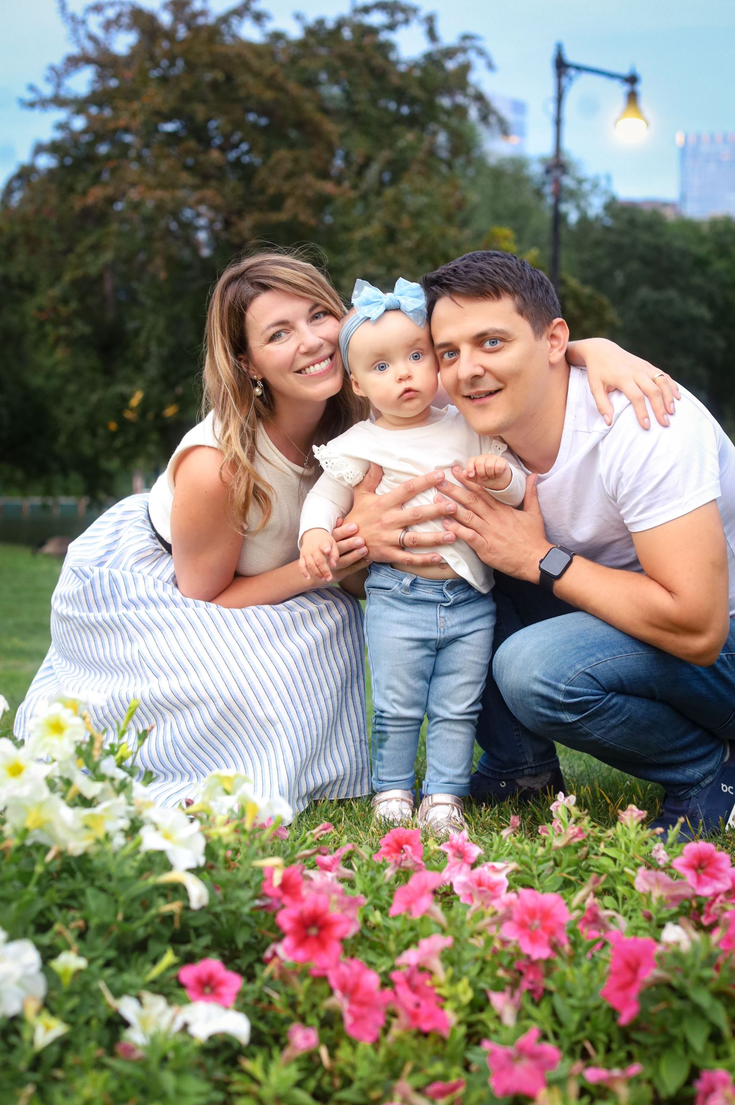 Young family with baby girl in blue bow posing near colorful flowers in a park.