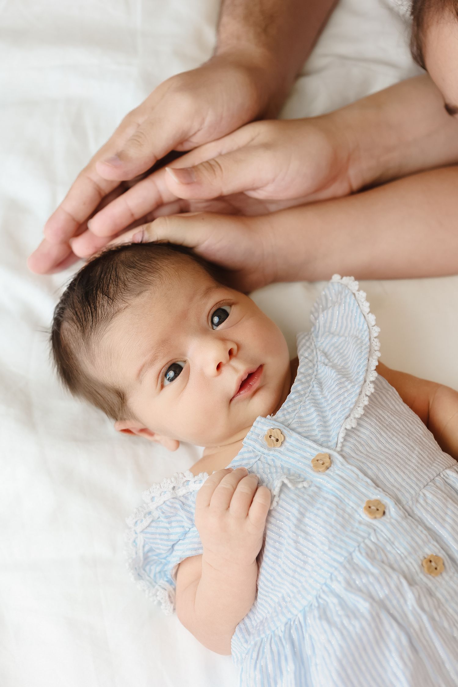 Alert newborn baby in blue striped dress lying on white bed with parents hands.