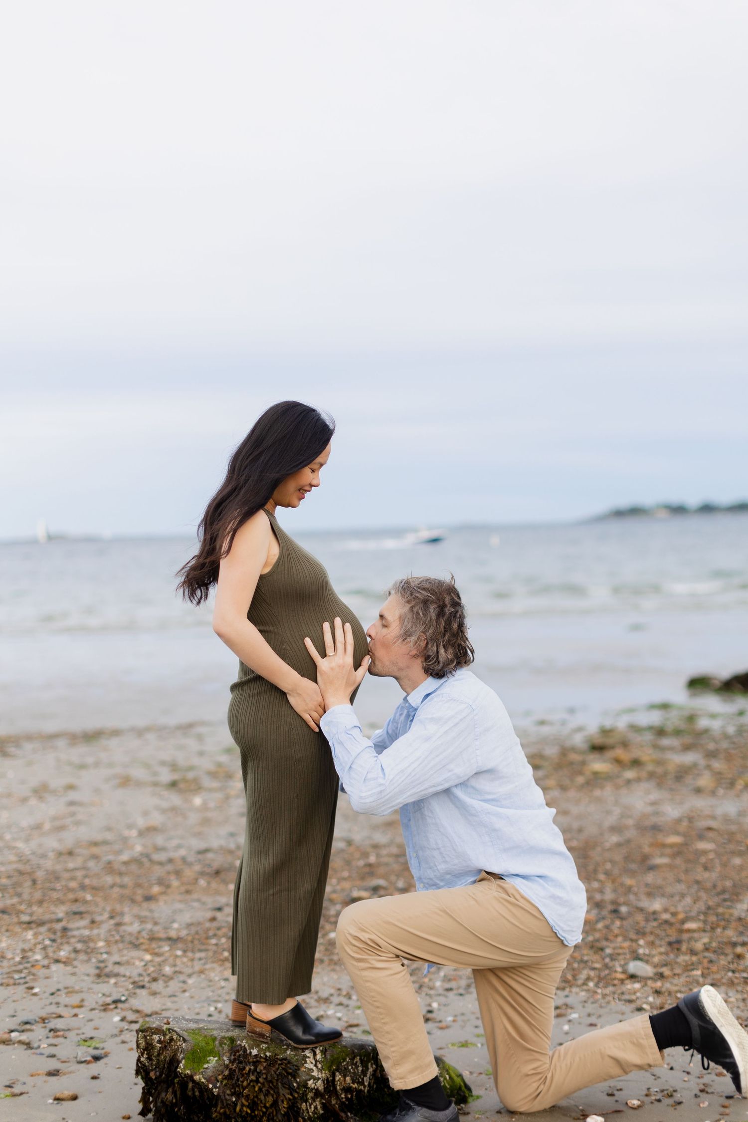Man kneeling on beach kissing pregnant woman's belly during maternity photoshoot by the ocean.