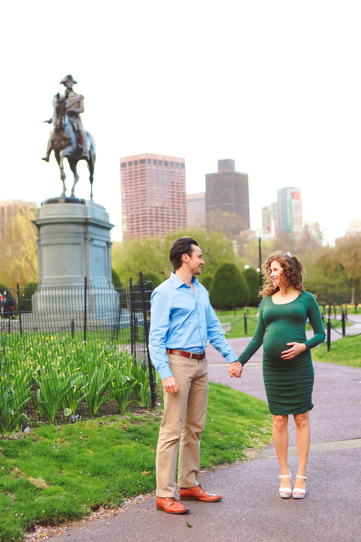 Pregnant couple holding hands in Boston Public Garden with George Washington statue and city skyline in background.