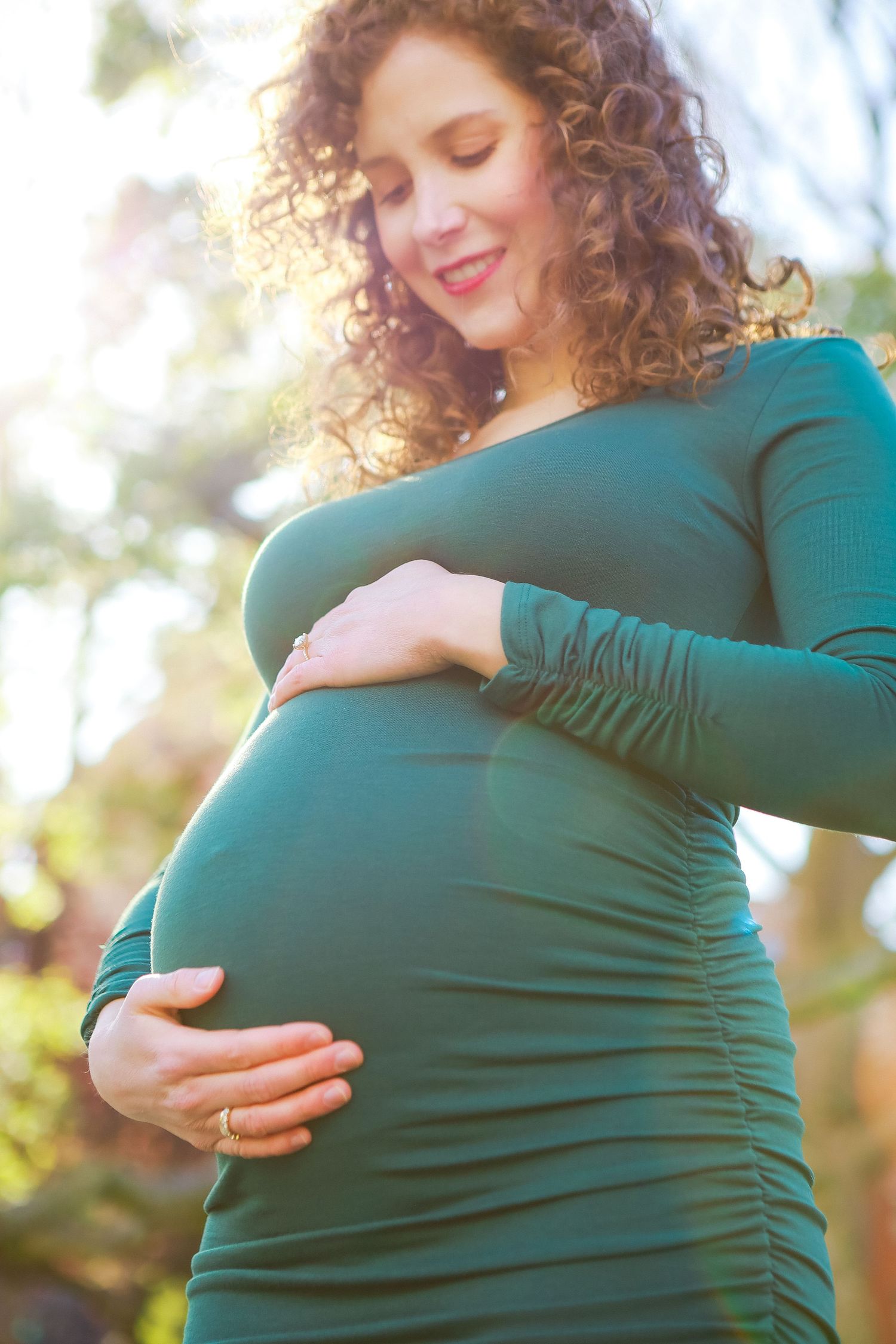 Curly-haired pregnant woman in teal dress cradles baby bump outdoors in warm sunlit natural setting.