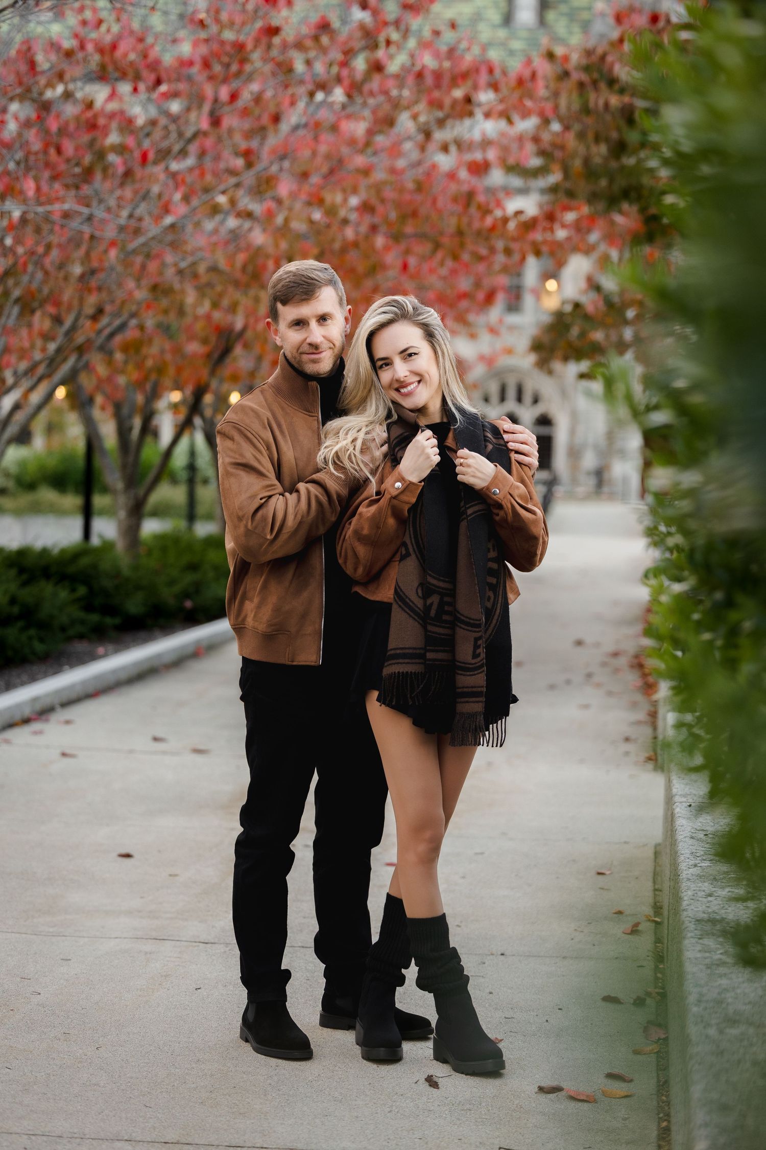 Smiling couple embracing on a pathway surrounded by red autumn foliage near a stone building.