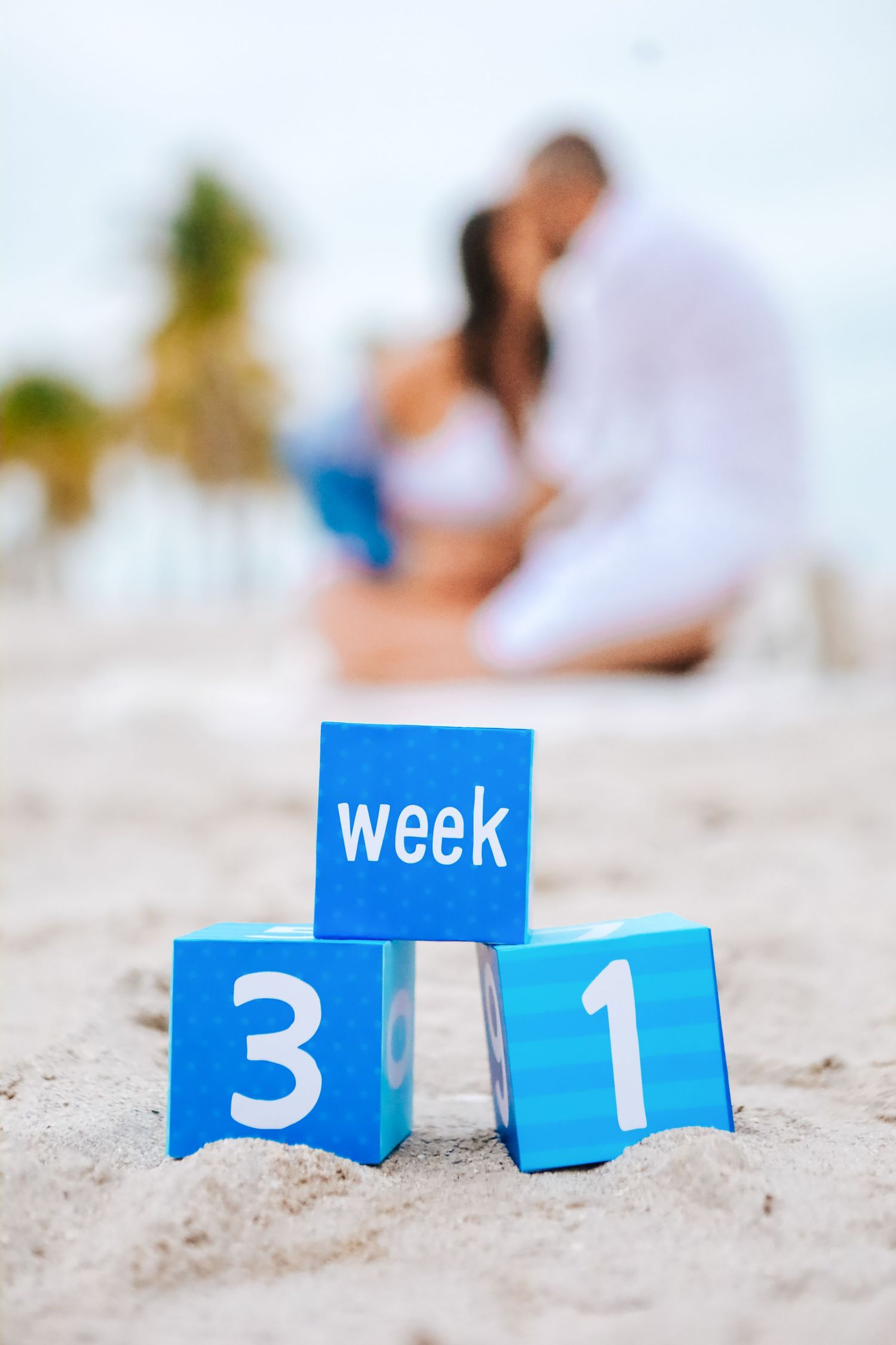 Blue blocks spelling week 31 in sand with kissing pregnant couple blurred in background on beach.