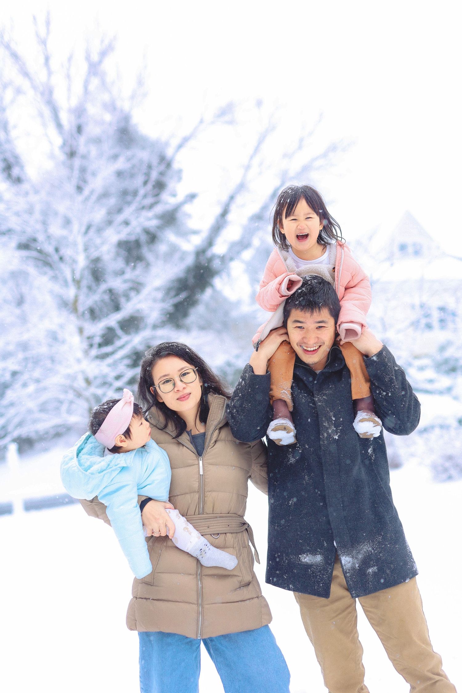 Happy Asian family of four playing in the snow, with children on parents shoulders in a winter wonderland.