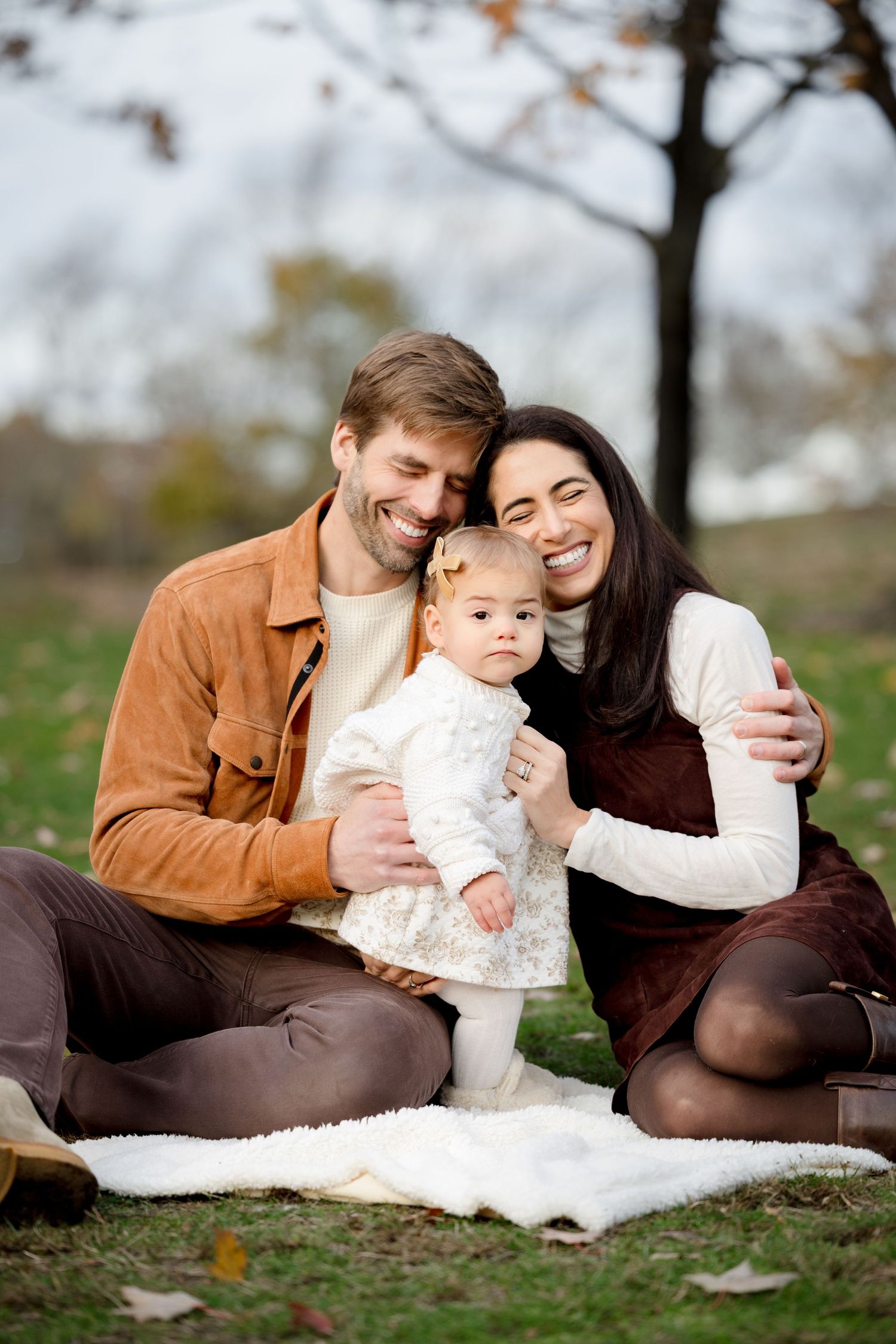 Smiling young couple sitting on a blanket in an autumn park with their standing toddler daughter.