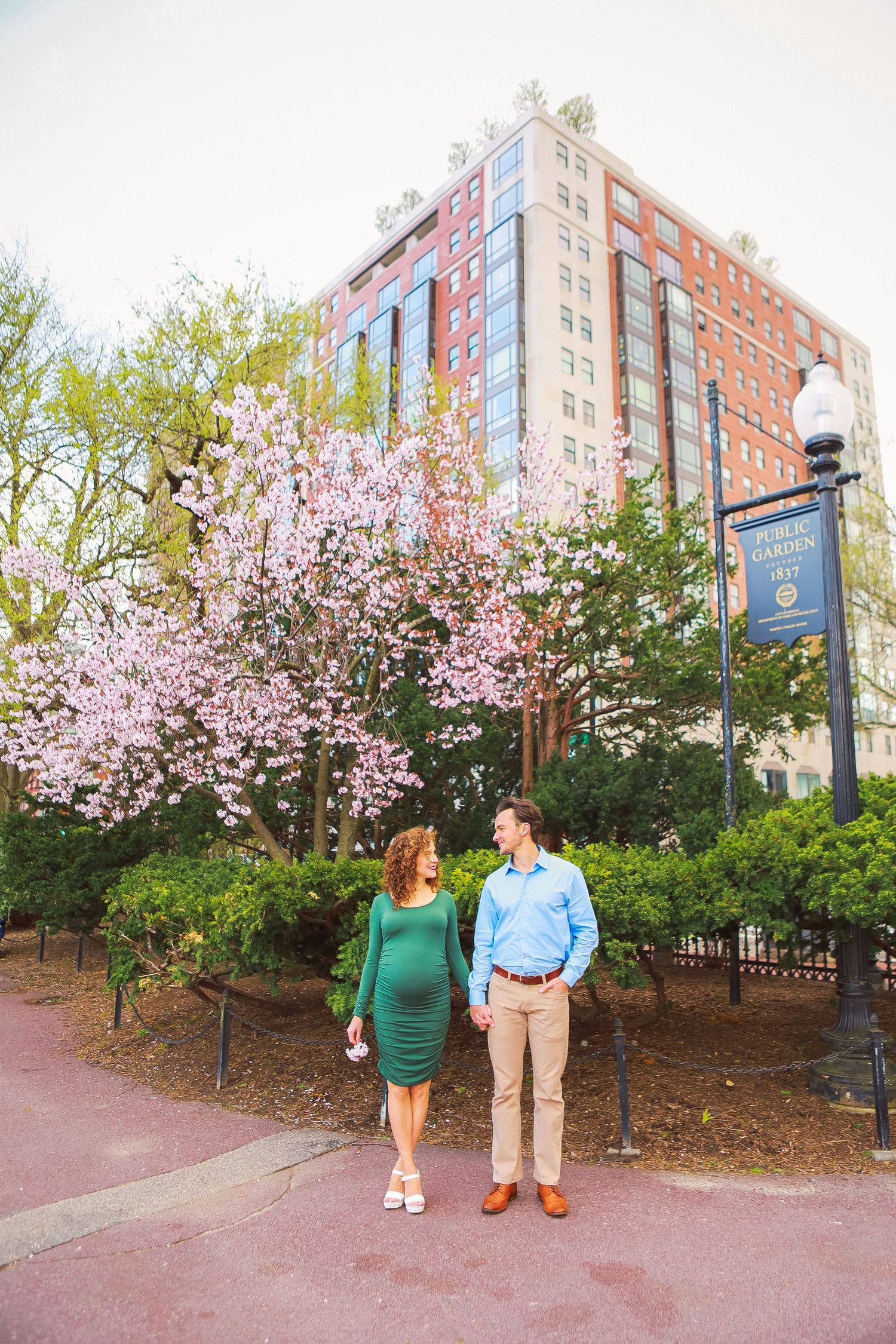 Pregnant couple holds hands and smiles at each other near blooming cherry blossom tree in urban park setting.
