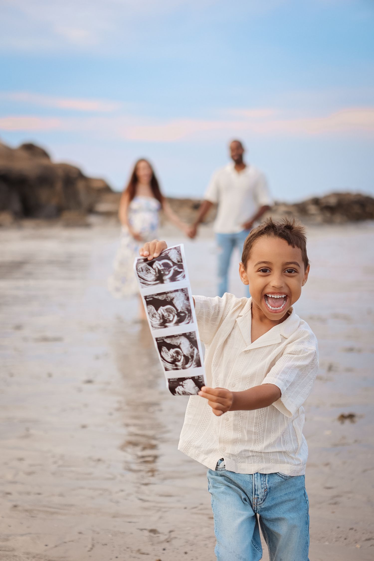 Excited child holding ultrasound photos on a beach with parents standing in the background.